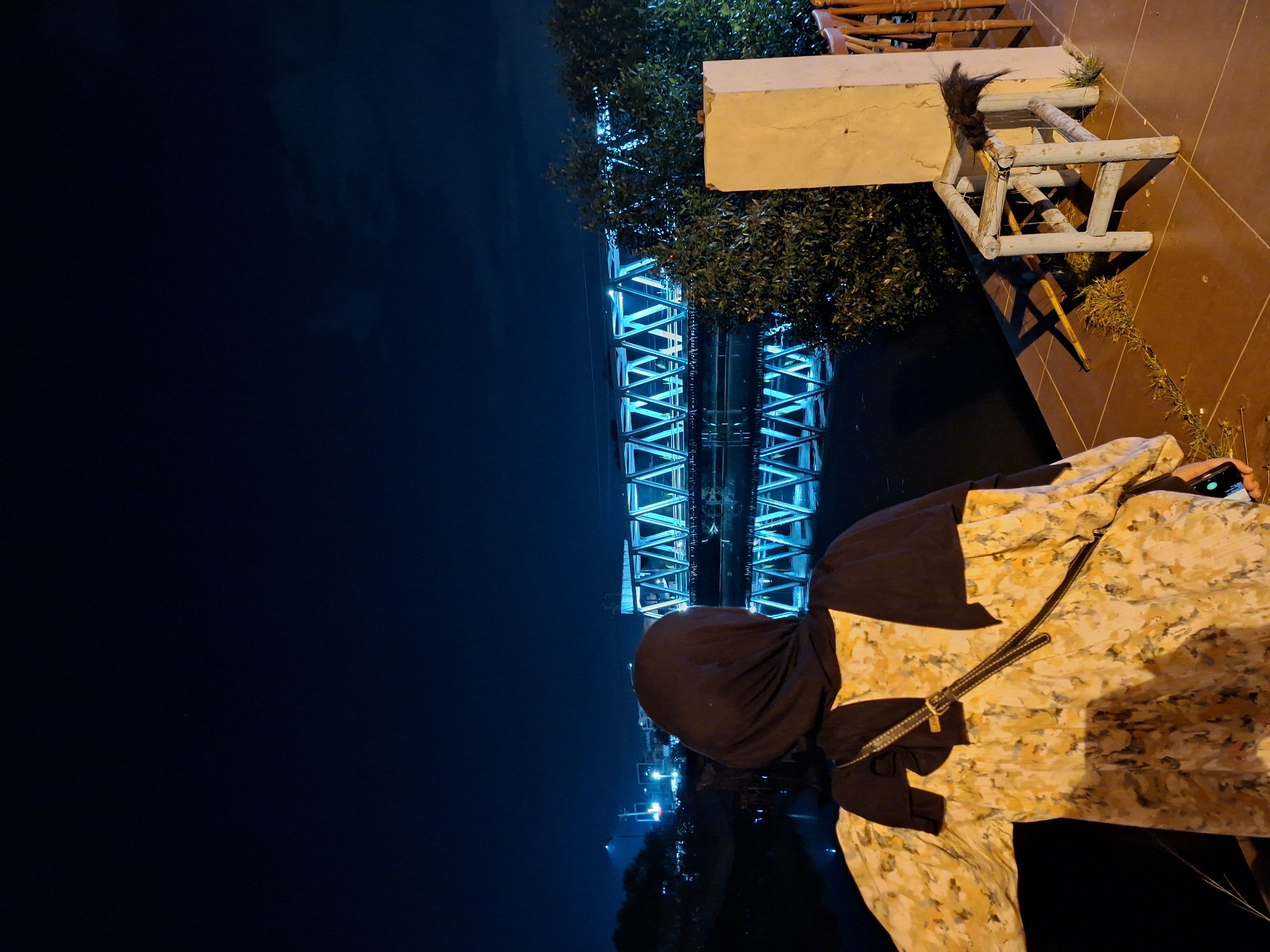 At night, a woman in a black head covering and floral patterned attire is standing next to a river at the Krueng Cunda River in Lhokseumawe, Indonesia. A blue-lit bridge is visible in the background, its lights reflecting on the water. Next to her is a low white wall and a weathered wooden chair.