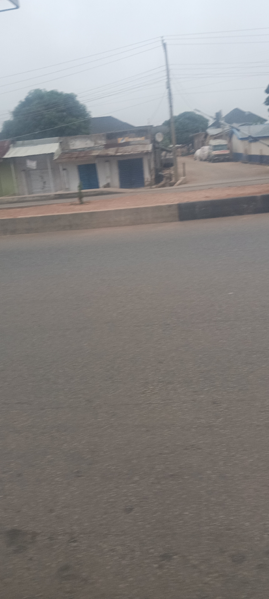 The image presents a static, eye-level street scene in a town or city, likely in Jos, Nigeria, as per the provided context. The foreground is dominated by a gray asphalt road, leading to a raised concrete median. This median has a reddish-brown top surface and a gray/black side facing the road, with a small, slender plant growing from it in the mid-left.
Beyond the median, a row of low-rise buildings lines the street under an overcast sky, suggesting a cloudy day or soft, diffuse lighting conditions. On the far left, a light-colored building with a white overhang is partially obscured by a large, dense green tree. Adjacent to it, a central building features a light-toned, somewhat weathered facade with two distinct blue roll-down shutters, characteristic of shop fronts. A faint, unreadable sign, possibly yellow and blue, can be vaguely discerned on this building's upper section.
Further to the right, additional structures with dark roofs are visible. A prominent utility pole stands on the right side of the frame, supporting numerous overhead wires that crisscross the sky. In the background on the right, a white or light-colored truck is parked, with another smaller vehicle possibly nearby. No people are visible in the scene, and no specific activity is occurring, conveying a quiet, everyday street ambiance.