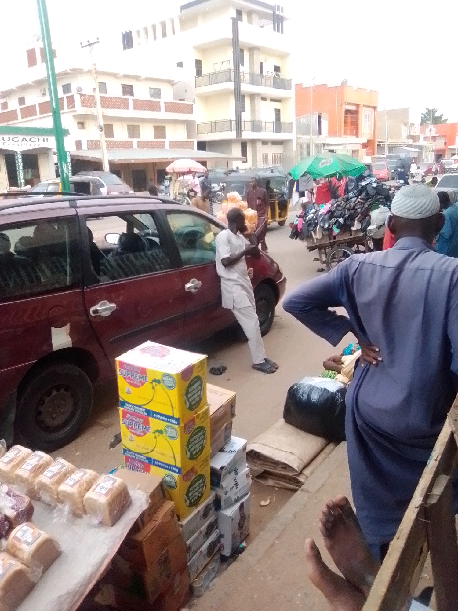 In the city of Arawa, Nigeria, a street scene unfolds with a vibrant marketplace atmosphere. The foreground features a display of bread loaves wrapped in plastic, arranged on a white surface, alongside stacks of boxed "Supreme Noodles" in bright yellow packaging. To the left, a maroon minivan is parked, its rear door slightly ajar. A man in a light-colored traditional outfit stands beside the minivan, holding a package. Further into the scene, a crowd gathers around a stall covered by a green umbrella, displaying an array of footwear. A rickshaw-style vehicle, a "Keke Napep," is visible to the left, with passengers. Buildings with multiple stories and balconies line the background, suggesting a bustling urban environment. People in various attire, including a man in a blue tunic and cap, are present, some interacting with vendors or observing the market. The ground appears to be paved, with debris scattered in places, indicative of an active commercial area.