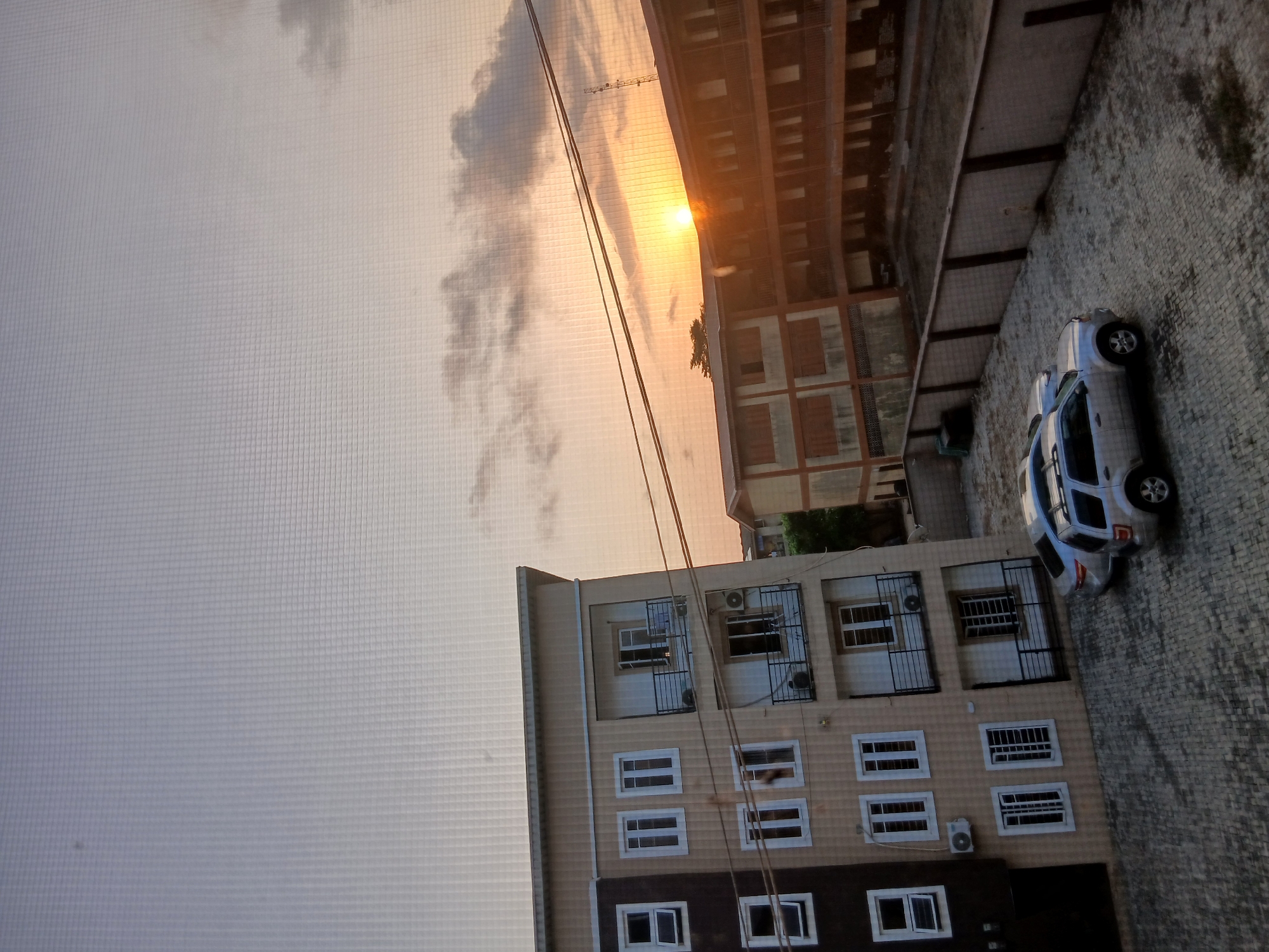The image captures a Lagos, Nigeria, urban scene during a sunset or sunrise. A multi-story building with light beige and dark brown facades featuring numerous windows and some with air conditioning units and security bars is the main structure. A silver-colored vehicle is parked adjacent to the building on a paved surface. Above, a sky with a gradient of colors from light gray to orange is visible, likely indicating a sunset or sunrise. Power lines are strung across the scene. The overall impression is of a residential or commercial area at dusk or dawn.