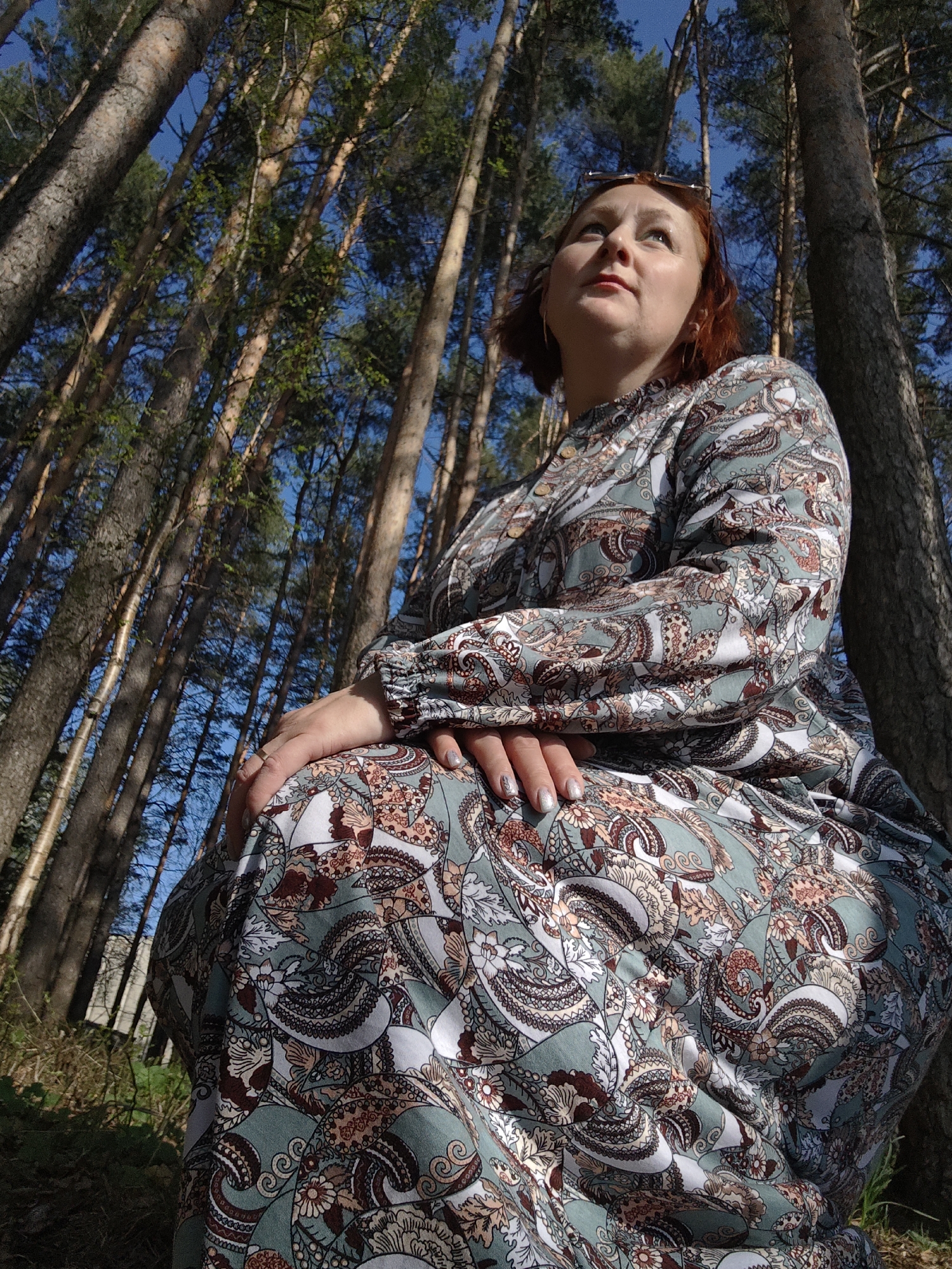 A medium-angle shot shows a woman with fair skin, red hair, and wearing a patterned dress sitting outside amongst tall trees. The camera is positioned low, looking up at the woman. The background consists of tall, thin trees with brown trunks that stretch upwards into a blue sky. Light filters through the trees, creating dappled shadows on the woman and the ground. The woman is looking up and slightly to her left. Her hands are clasped in her lap. The location is Chaikovsky, Russia.