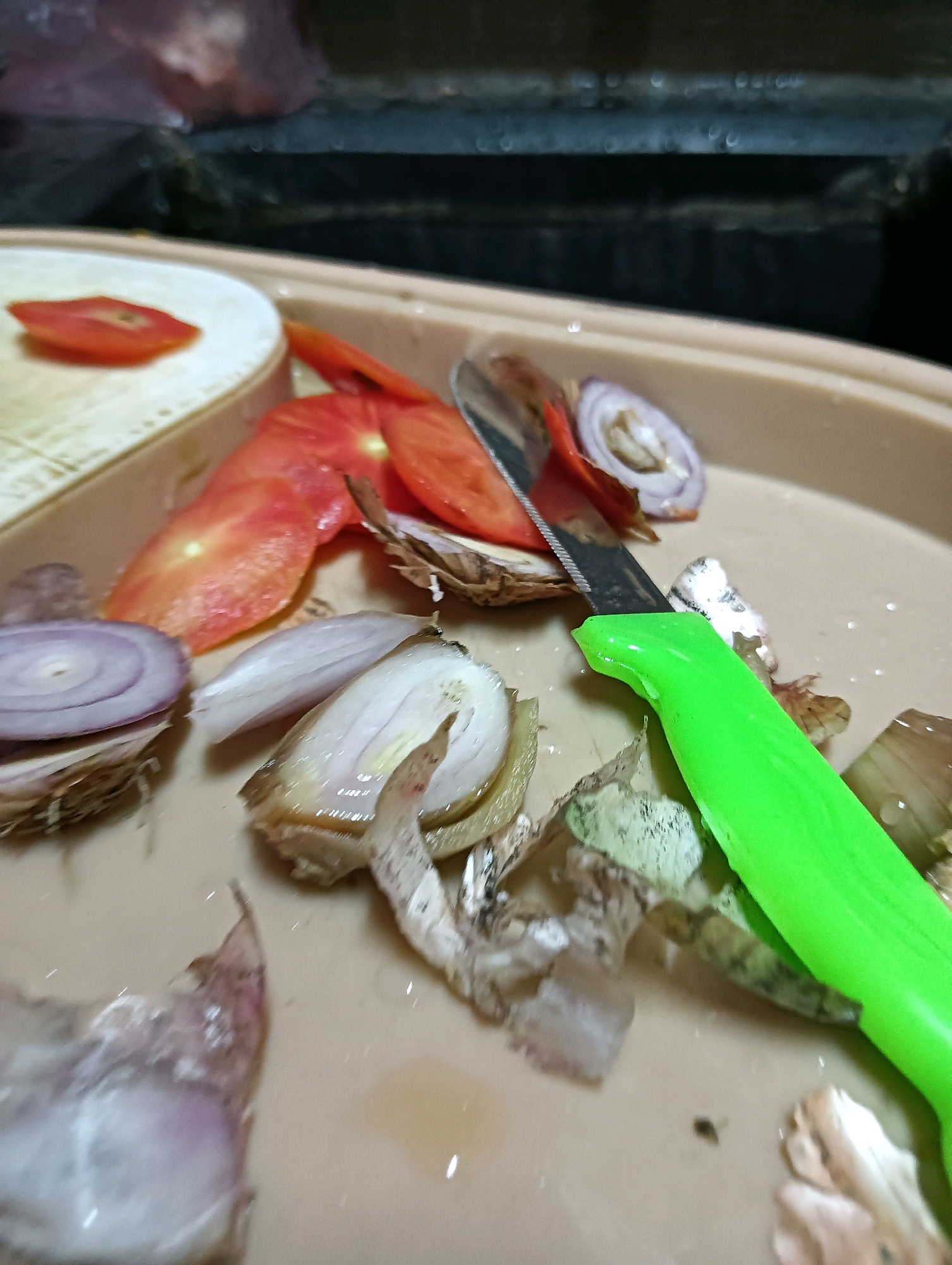 In Hosapete, India, a close-up, high-angle shot captures a light brown, plastic tray on a dark countertop. The tray contains various food preparation remnants. To the left, a slice of tomato rests on a white, ridged surface. Next to it, several red tomato slices and a portion of a red onion are scattered. A knife with a serrated blade and a bright green handle is positioned diagonally across the tray, its tip touching the red onion. Other items in the tray include discarded onion skins, some peeled outer layers of shallots, and a few small, wet, brown flecks. The overall scene suggests the process of preparing vegetables, likely for a meal.