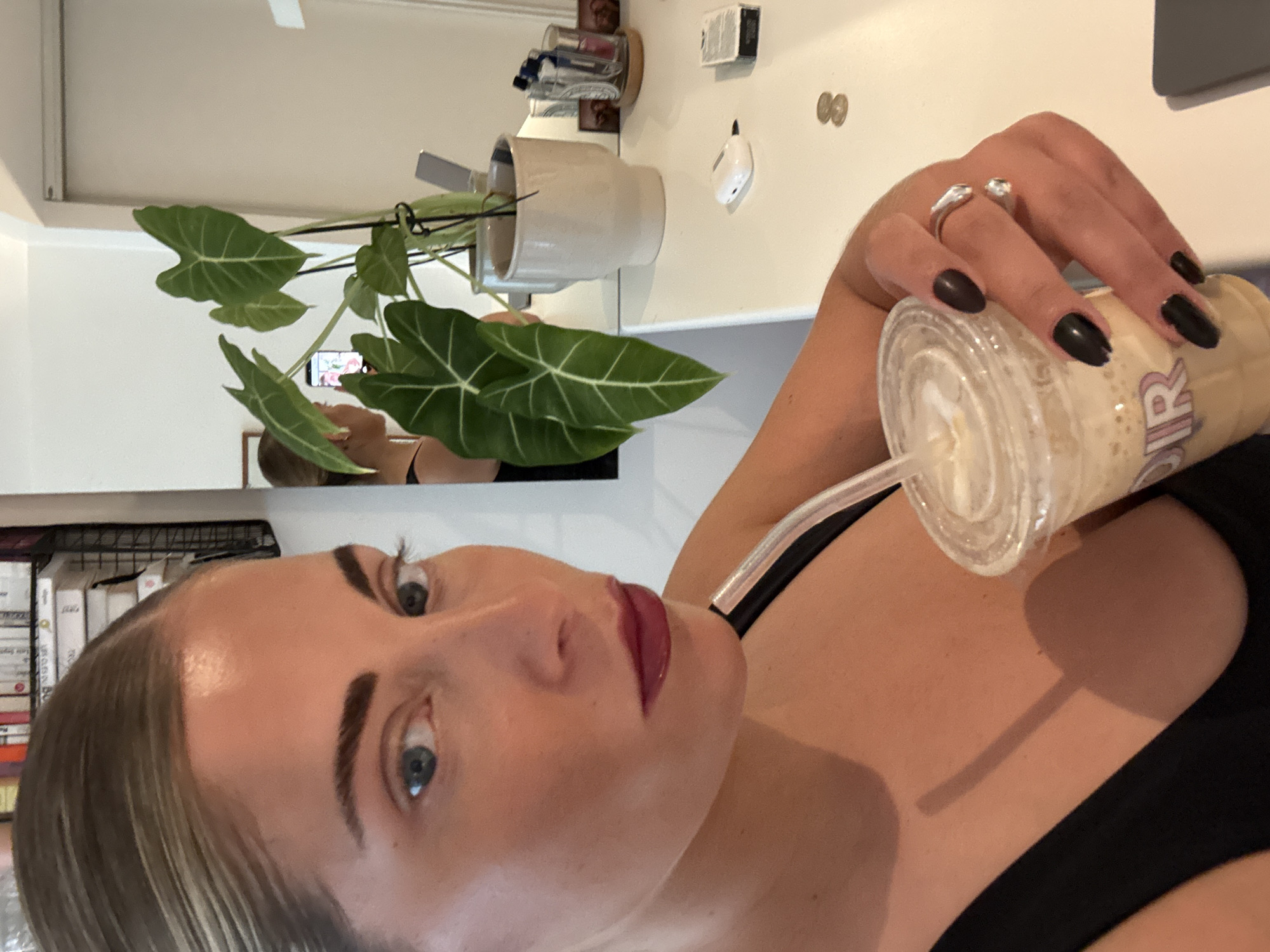 A woman with light skin, light brown and blonde hair, and blue eyes is holding a beverage in a clear plastic cup with a straw. She is wearing a black top and has dark nails and dark red lipstick. A silver ring is visible on her hand. In the background, there is a potted plant on a surface. A mirror reflects the woman taking the photo, holding a phone. Various items are on the surface, including coins and a small white object. The image appears to be taken indoors. The location in the city of Paris, France, is not verifiable from the image.