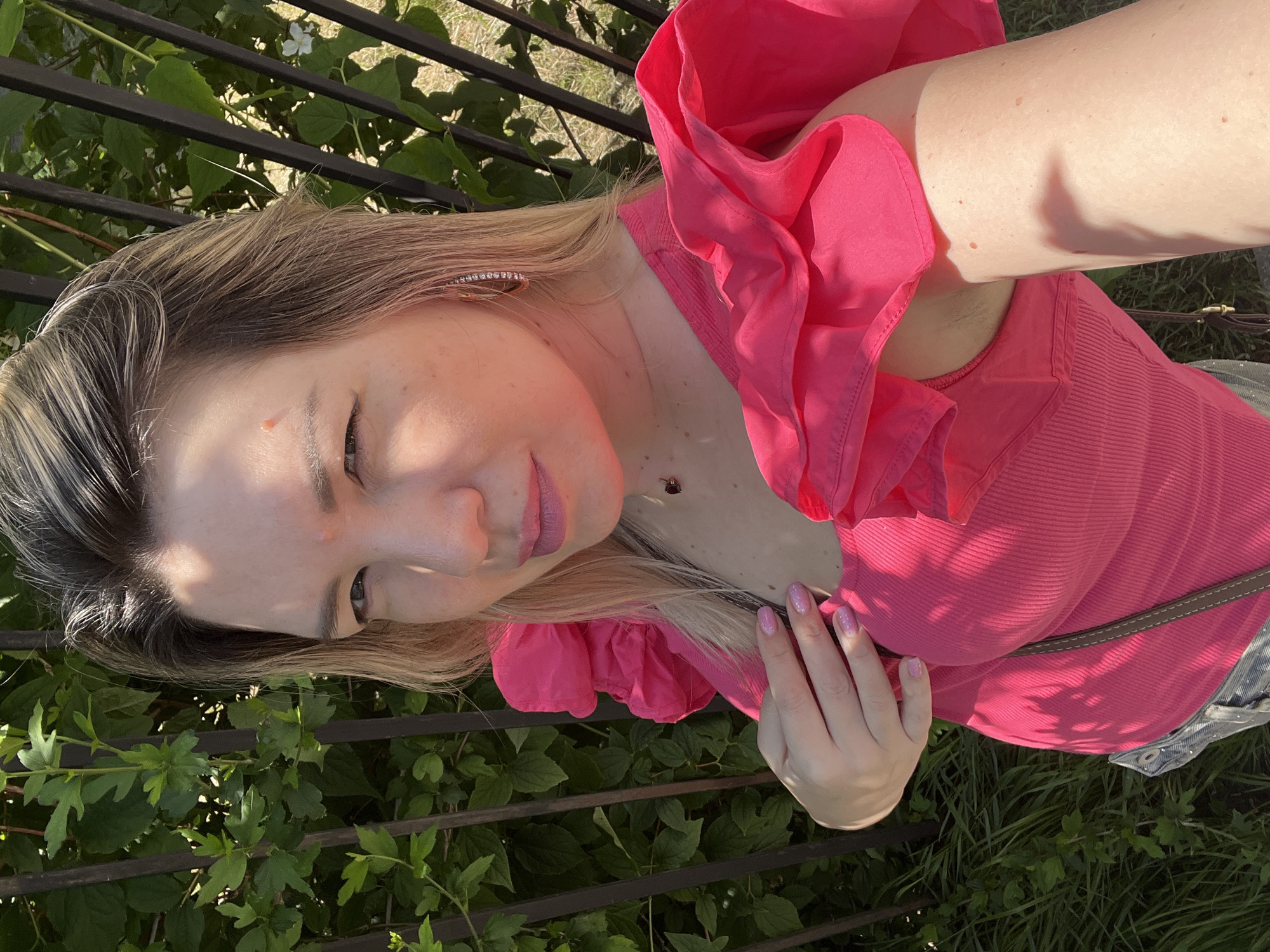 An adult female, characterized by fair skin and light-colored hair, is depicted in a close-up, selfie-style photograph taken outdoors in the city of Sochi, Russia. She wears a vibrant fuchsia-pink top featuring a ribbed bodice and prominent ruffled sleeves. A small, reddish raised mark is visible on her forehead, positioned above her right eyebrow. Her left eye is partially closed, indicative of bright sunlight, while her lips are colored with pink lipstick and slightly parted. A delicate necklace with a dark heart-shaped pendant is worn around her neck, and a sparkling hoop earring is visible in her right ear. Her fingernails are painted with pink glitter polish. Her left hand is placed on her chest. The background consists of dense green foliage behind a dark, vertical-barred metal fence, with patches of grass visible through and below the greenery. The scene is brightly illuminated by direct sunlight, casting shadows on the subject and the immediate environment.