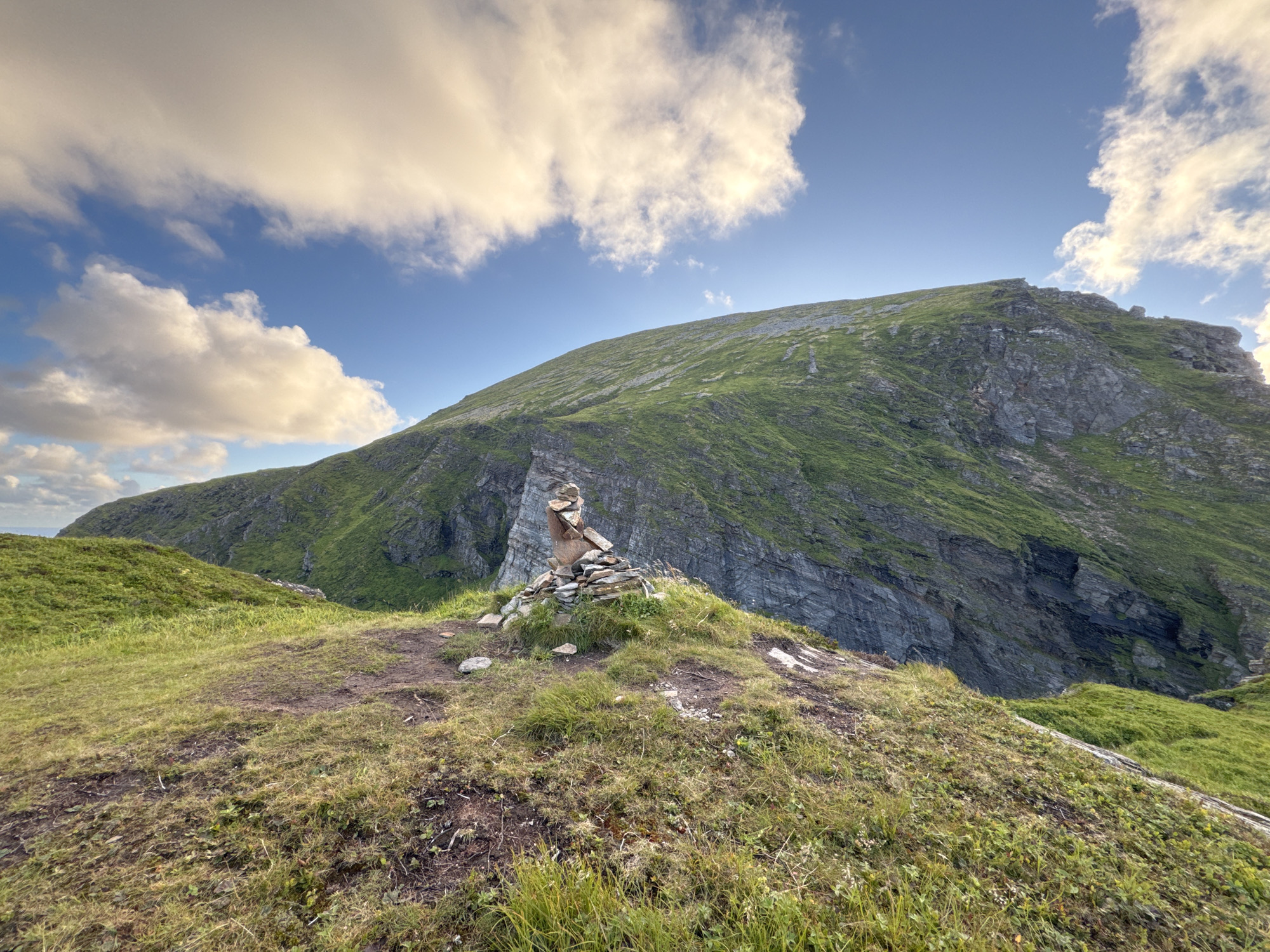 This is a daytime, wide-angle, eye-level shot of a grassy hilltop with a cairn in the foreground. The cairn is a stack of stones, the largest of which is a rough-hewn reddish-brown stone at the top. The hill slopes gently upward to the right, meeting a steep, rocky mountainside covered in green grass. The mountainside recedes into the distance, forming a large, imposing peak against a blue sky with scattered white clouds. The environment appears to be a remote, natural landscape, likely in Lovund, Norway, given the mention of its location. No people are visible in the image. The overall impression is one of rugged natural beauty and tranquility.