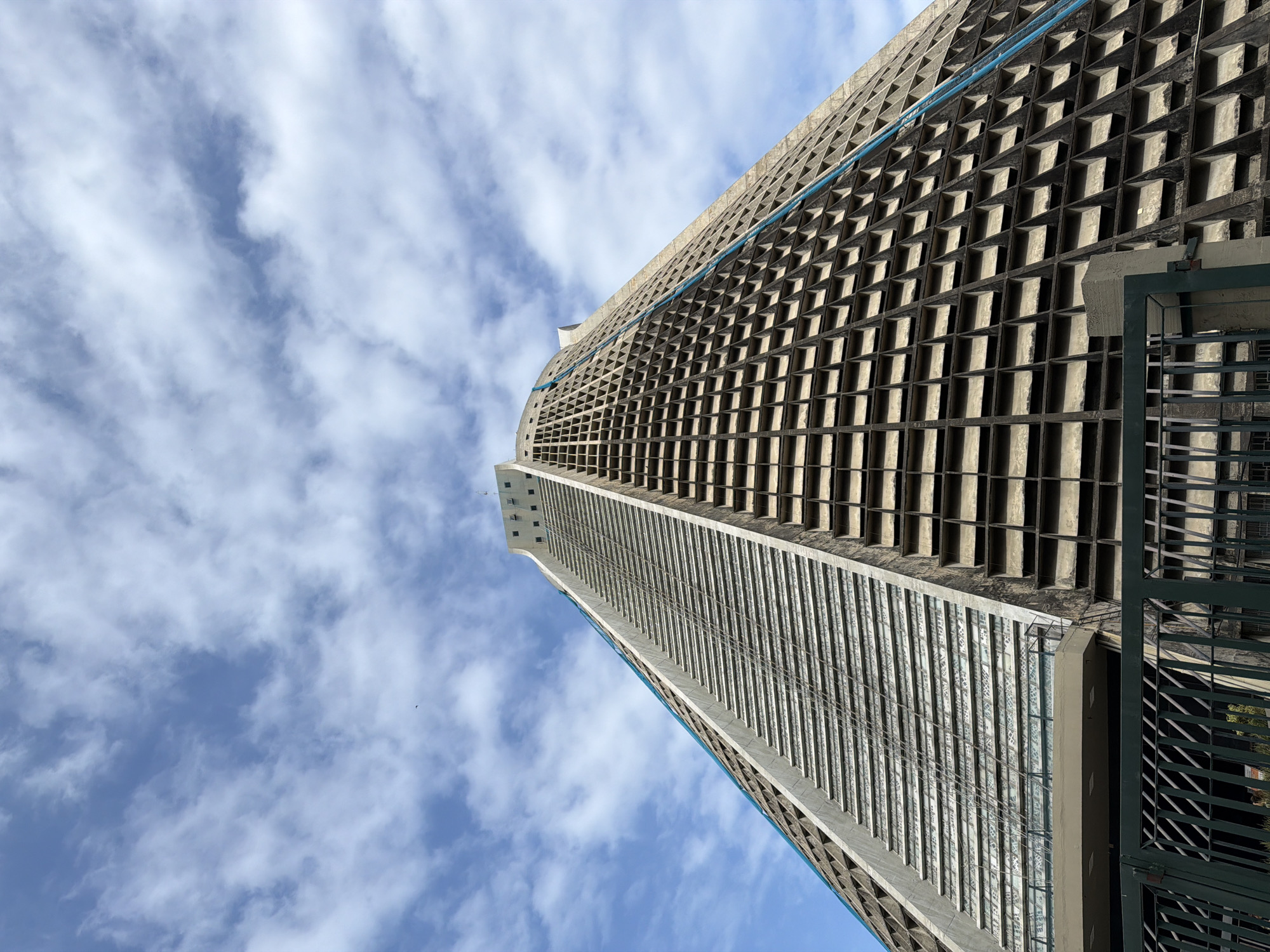 The image shows a low-angle view of the Metropolitan Cathedral of Saint Sebastian in Rio de Janeiro, Brazil, under a partly cloudy blue sky. The cathedral is a modern, conical-shaped building constructed from concrete. Its facade features a steep, tiered design with rows of geometric openings and a prominent, sloped section with horizontal grooved panels. A thin blue line runs diagonally along the side of the building, marking a seam or decorative element. In the foreground, a dark green metal fence partially obscures the base of the structure, with some greenery visible behind it. The overall impression is one of imposing architecture against a natural sky.