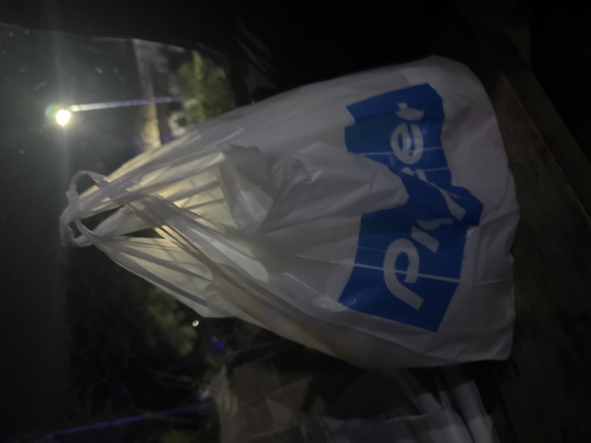 This is a close-up, low-light photograph of a white plastic shopping bag with blue lettering on its side. The bag appears to be full and is held by its handles. The blue lettering is partially visible and appears to spell out "Price". The bag is the primary subject, positioned in the foreground and taking up most of the frame. The background is dark and out of focus, with hints of greenery and what appears to be a street light with a noticeable lens flare. The overall impression is that the photograph was taken at night, possibly in an outdoor setting given the faint lighting and what looks like trees in the background. There are no people visible in the frame. The scene is generic, with no strong location cues beyond the mention of Abuja, Nigeria. The image does not convey any specific emotion or action, other than the simple presentation of a shopping bag.