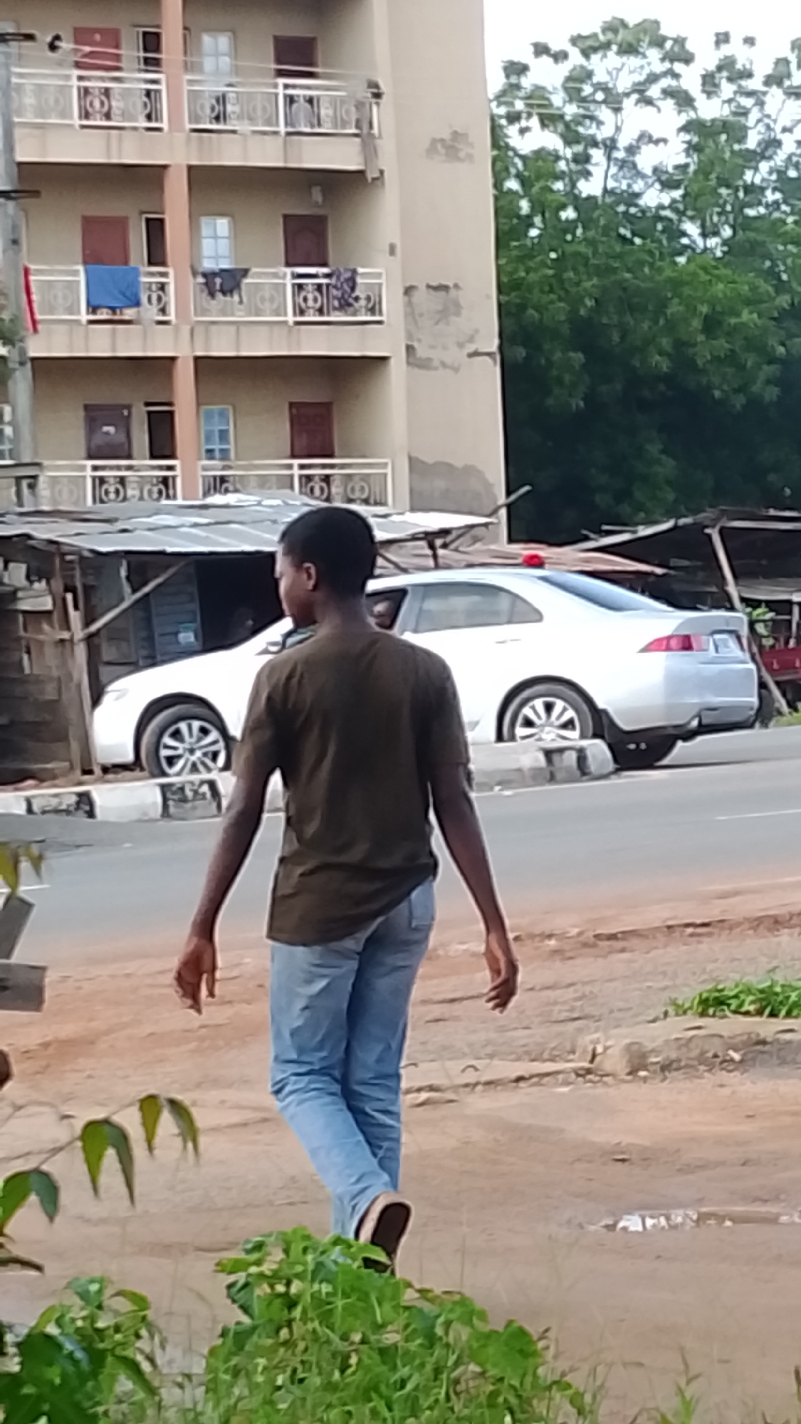 A young Black male wearing a dark olive green t-shirt and light blue jeans walks away from the camera, towards the right of the frame. He is walking on a dirt path adjacent to a paved road. In the background, a white sedan car is parked on the side of the road, behind a makeshift stall constructed of corrugated metal and wood. Beyond the car and stall, a multi-story building with balconies can be seen. The building is light brown with visible signs of weathering and peeling paint. Green trees with dense foliage are visible behind the building. The sky is overcast. The scene appears to be in Adankolo, Nigeria.