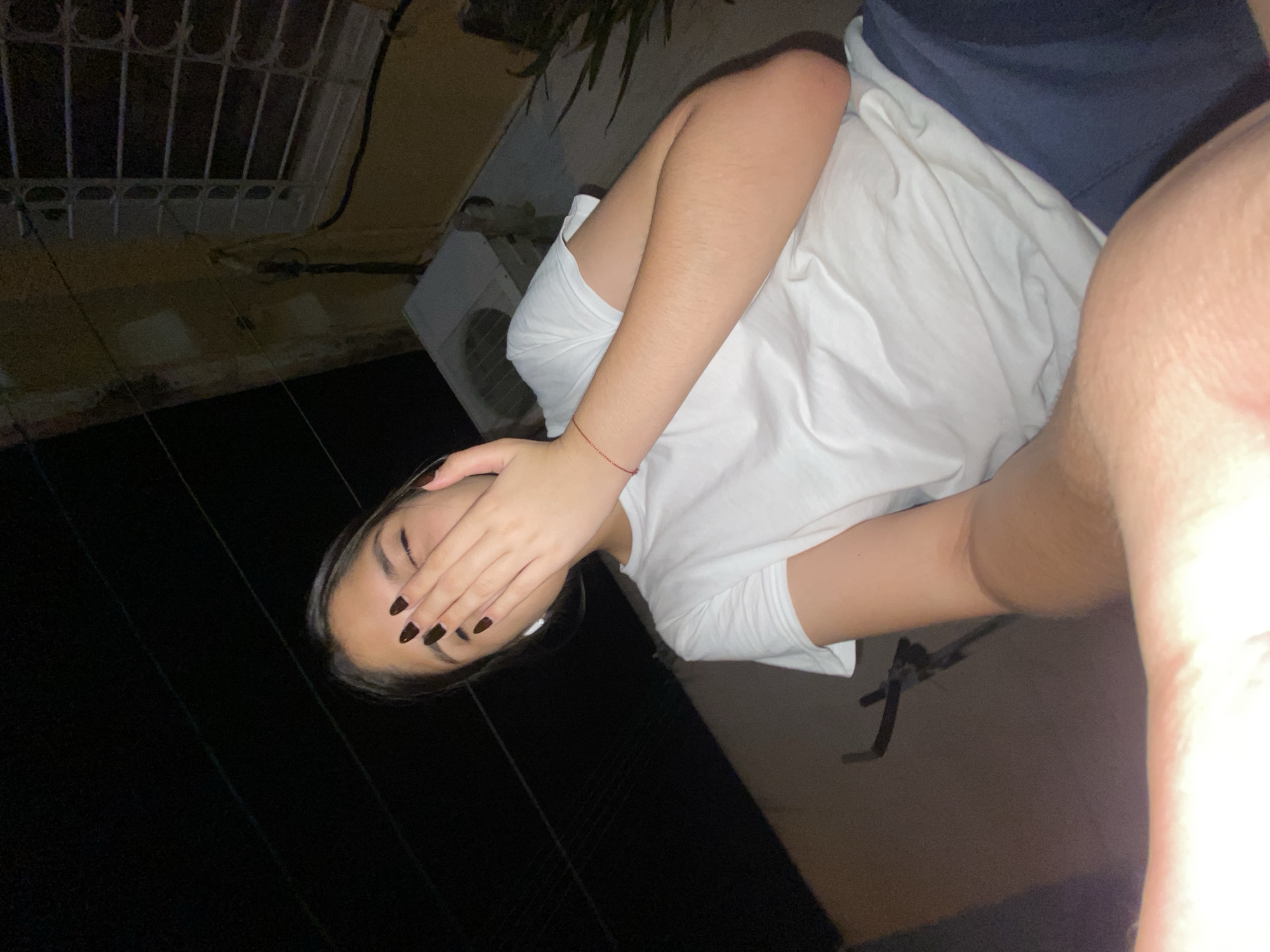 The image shows a person with dark hair and dark nail polish covering their eyes with their hand. They are wearing a white t-shirt and appear to be lying down. The background includes what looks like an exterior building with a window and an air conditioning unit. The scene is dark, with some visible architectural elements. There's not enough information to reliably place the location in Cotoperíz, Venezuela.