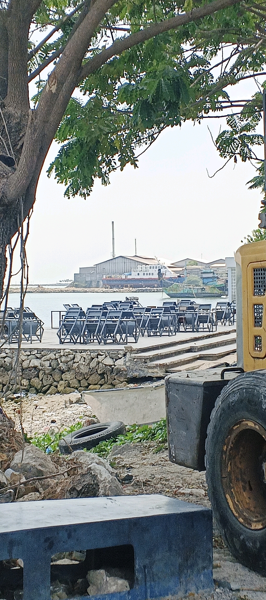 In Kandangsemangkon, Indonesia, this outdoor scene captures a waterfront area under a bright, overcast sky. In the foreground, a large, dark blue concrete structure with a rectangular opening sits partially in frame, obscuring the ground which consists of gravel and scattered debris, including a deflated tire and green vegetation. To the right, the large yellow machinery of a forklift or similar construction vehicle is visible, with a prominent tire and part of its chassis showing. Behind this, a grey stone wall lines the edge of a raised concrete walkway.
The walkway is occupied by numerous identical black chairs, stacked and arranged in neat rows, suggesting an outdoor seating area that is not currently in use. Beyond the chairs, a body of water is visible, with several boats and a larger vessel docked near an industrial building. This building, with its large roof and tall chimney, appears to be a factory or processing plant. Another, smaller, multi-colored boat is also moored on the water.
The background is dominated by the pale sky and the distant industrial structures. A large tree with thick branches and green foliage extends into the upper left quadrant of the image, framing the view of the water and the industrial complex. The overall impression is one of a working port or industrial waterfront, with elements of disuse and preparation for an unknown activity.