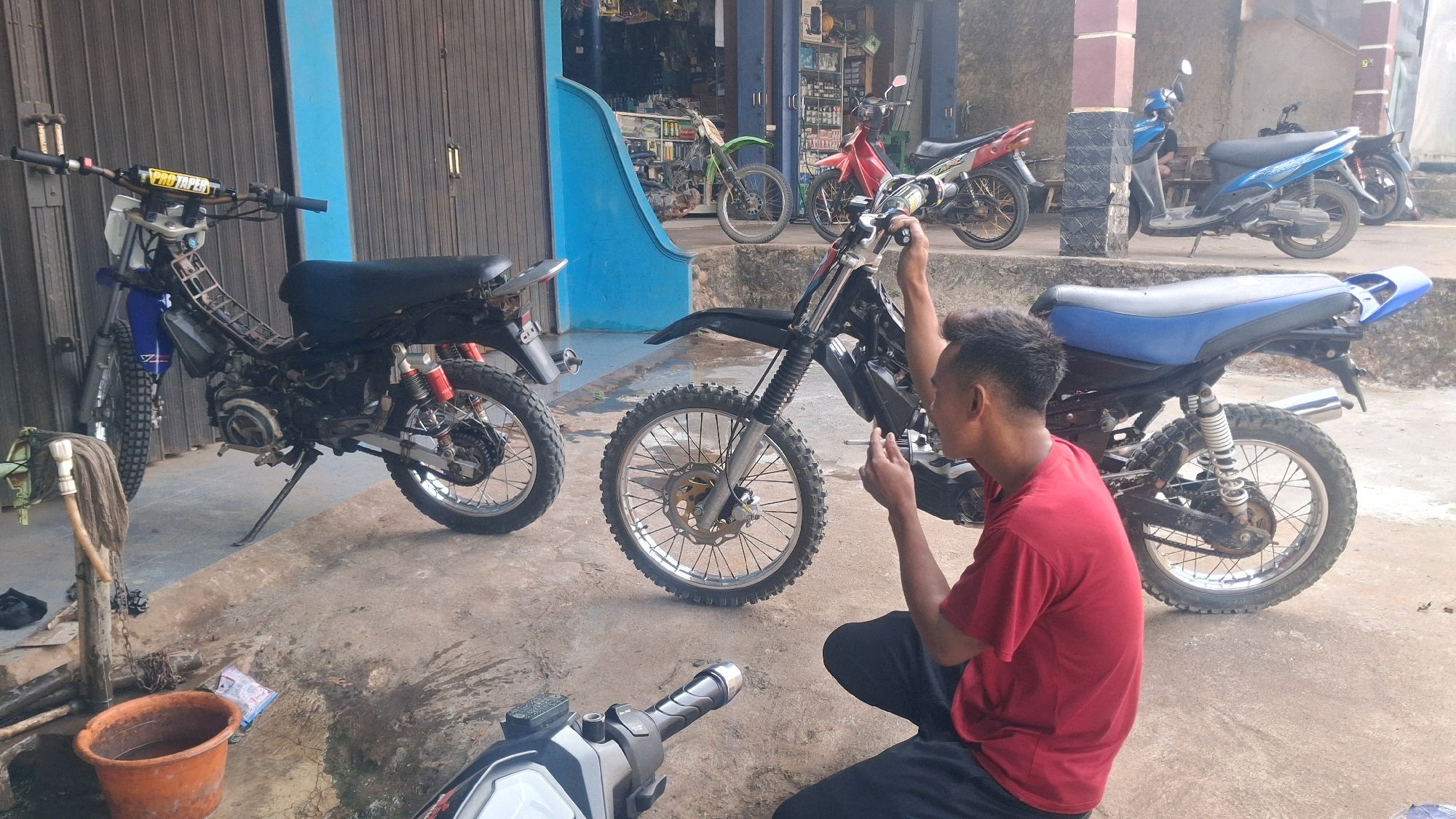In Karang Sari, Indonesia, a man in a red t-shirt and dark pants crouches next to a disassembled dirt bike. The bike, with its blue seat and chrome exhaust pipe, is partially stripped down, revealing its internal components. In the background, other motorcycles are parked outside a building that appears to be a shop, with shelves stocked with goods visible through an open doorway. A smaller, blue scooter is also present, along with other motorcycles in varying states of repair or storage. The ground is a concrete surface, showing signs of use and some debris. The scene suggests a casual repair or maintenance area adjacent to a commercial establishment.