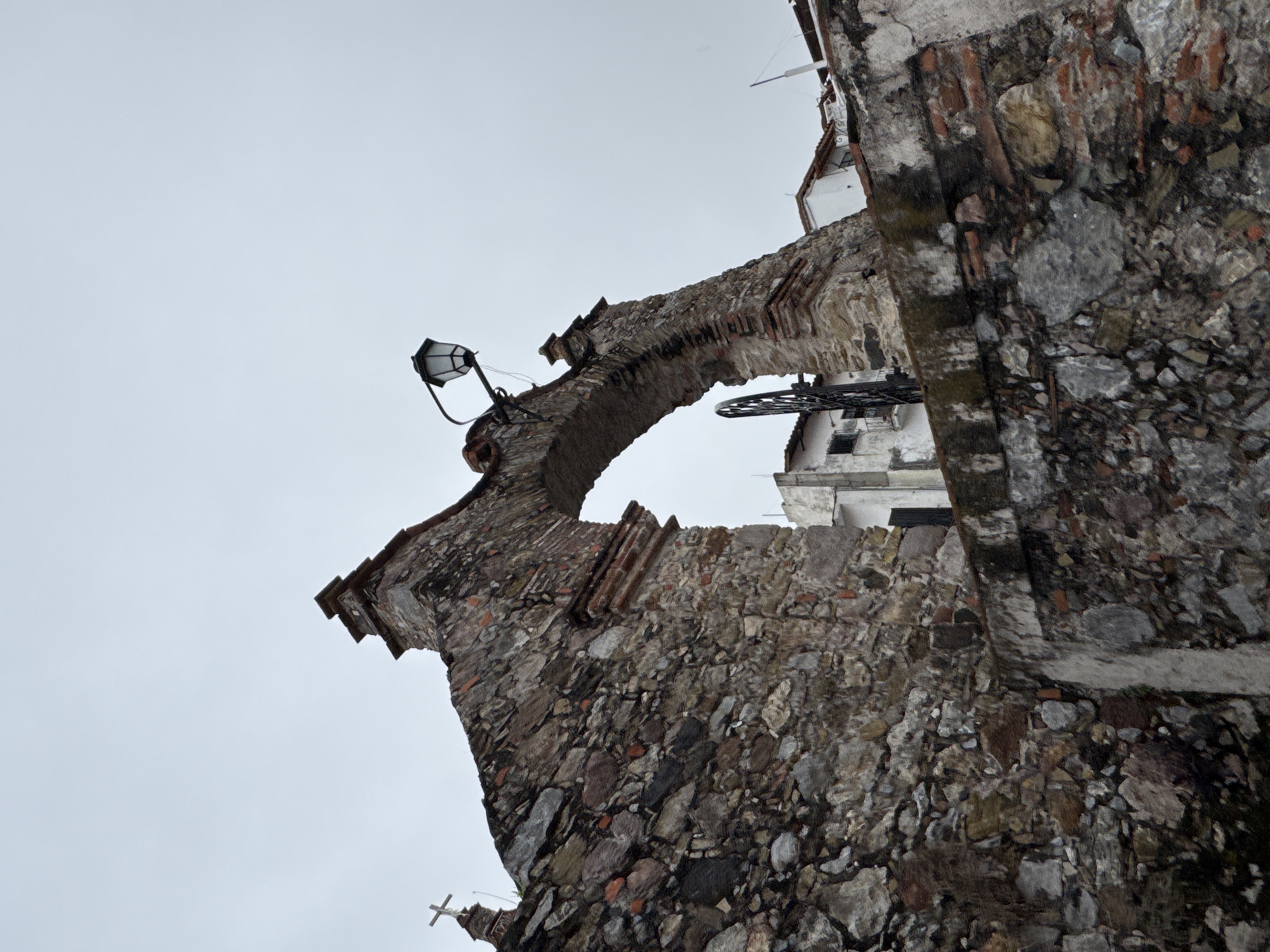 The image depicts a stone archway, likely part of a historical structure in Taxco, Mexico, given its rugged, rustic construction with visible stones and bricks. A cross is visible atop a turret on the left side of the arch. A traditional-style lantern is mounted on the archway itself. In the background, glimpses of white buildings with red-tiled roofs are visible, suggesting an urban or town setting, consistent with Taxco's architecture. The sky is overcast, creating a diffused light across the scene. The perspective is low-angle, looking up at the archway.