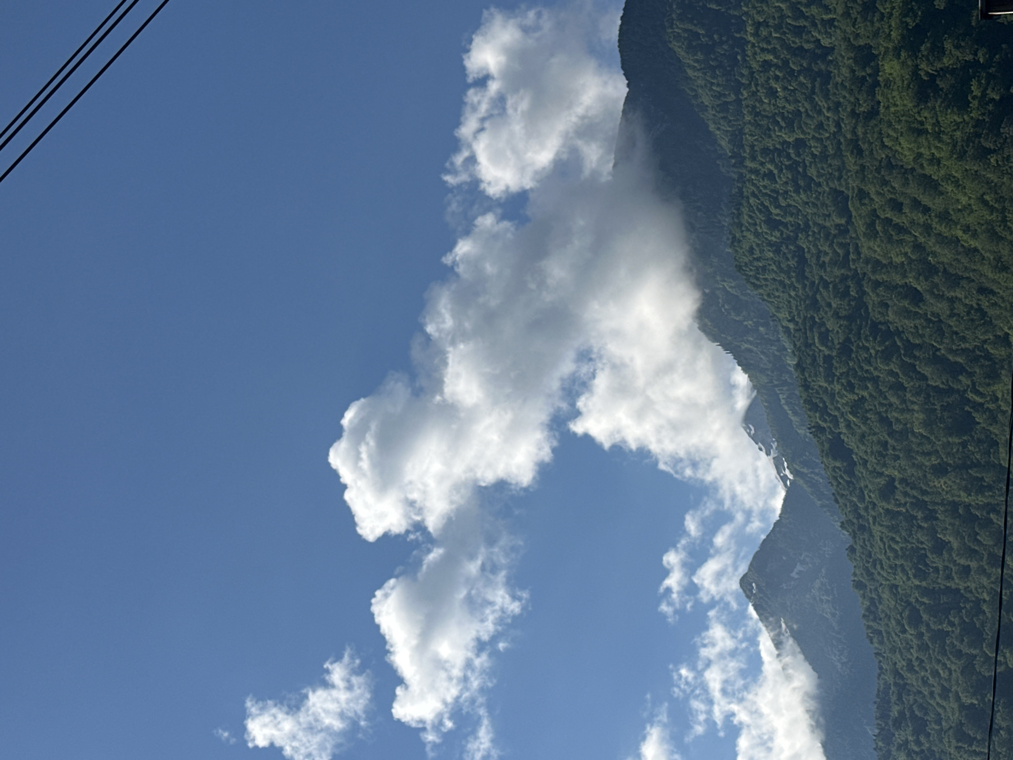The image captures a view of the mountains near Krasnaya Polyana, Russia, under a bright blue sky. Lush green, densely forested slopes dominate the lower half of the frame, rising towards jagged peaks. Patches of snow are visible on the higher elevations of some mountains. White, fluffy cumulus clouds are scattered across the sky, with some clouds partially obscuring the mountain peaks, creating a misty effect. Two black power lines are diagonally visible in the upper right corner of the image. There are no people or animals visible in the scene. The lighting suggests it is daytime, with the sun illuminating the clouds and the mountains.