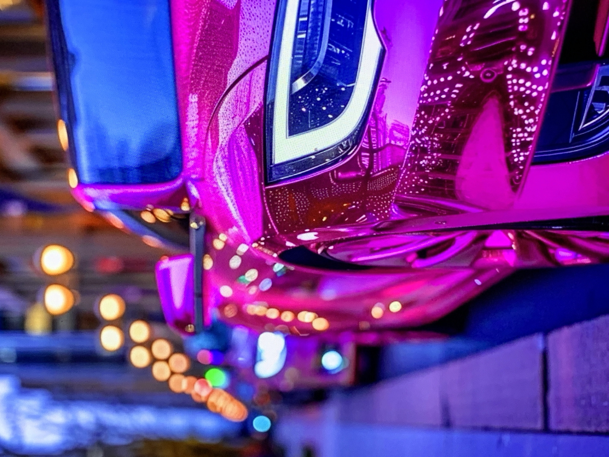 A close-up, side-view shot of a shiny, magenta-colored car is captured at night in an urban setting. The car's headlight, a sleek, angular design with bright white LED accents, is prominently featured on the right side of the frame. Reflections of city lights, appearing as numerous small, bright dots, shimmer across the car's polished surface. In the background, out-of-focus streetlights with a warm, orange glow line the street, and the blurred shapes of other vehicles and buildings suggest a city environment. The overall lighting is atmospheric, with a strong presence of purple and blue hues, creating a neon-like effect.
