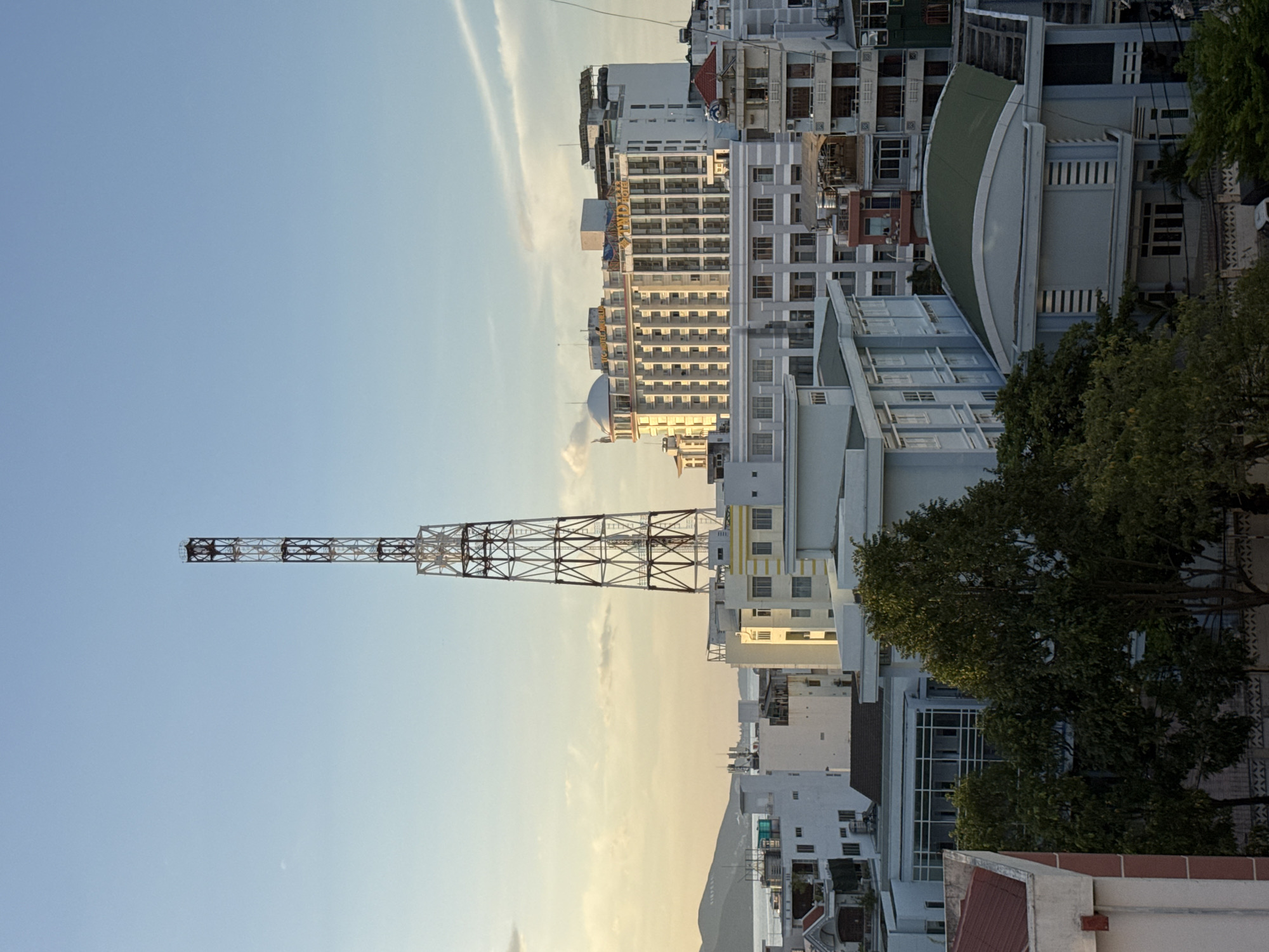 The image depicts a cityscape of Nha Trang, Vietnam, under a clear sky with soft, diffused sunlight. Dominating the skyline is a tall, red and white lattice tower, likely a telecommunications or broadcasting mast, positioned centrally in the upper half of the frame.
In front of and surrounding the tower are various buildings. A prominent, large white hotel with many windows, a rounded section topped by a dome, and signage that reads "YND HOTEL" is visible to the right of the tower. Other multi-story buildings, varying in color from white and yellow to grey and beige, fill the middle and lower portions of the image. Some of these structures appear to be residential or commercial buildings, with numerous windows and balconies.
The foreground is occupied by lush green trees, their branches extending upwards and obscuring parts of the lower buildings. A portion of a building with a red roof is visible on the far left. In the distance, a body of water, likely the sea, can be seen beyond the city's buildings, with hills or mountains forming a backdrop. The overall impression is a bright, sunny day in an urban coastal environment. There are no people or animals visible in the scene. The lighting suggests it is either early morning or late afternoon, given the warm tones in the sky and on the buildings.