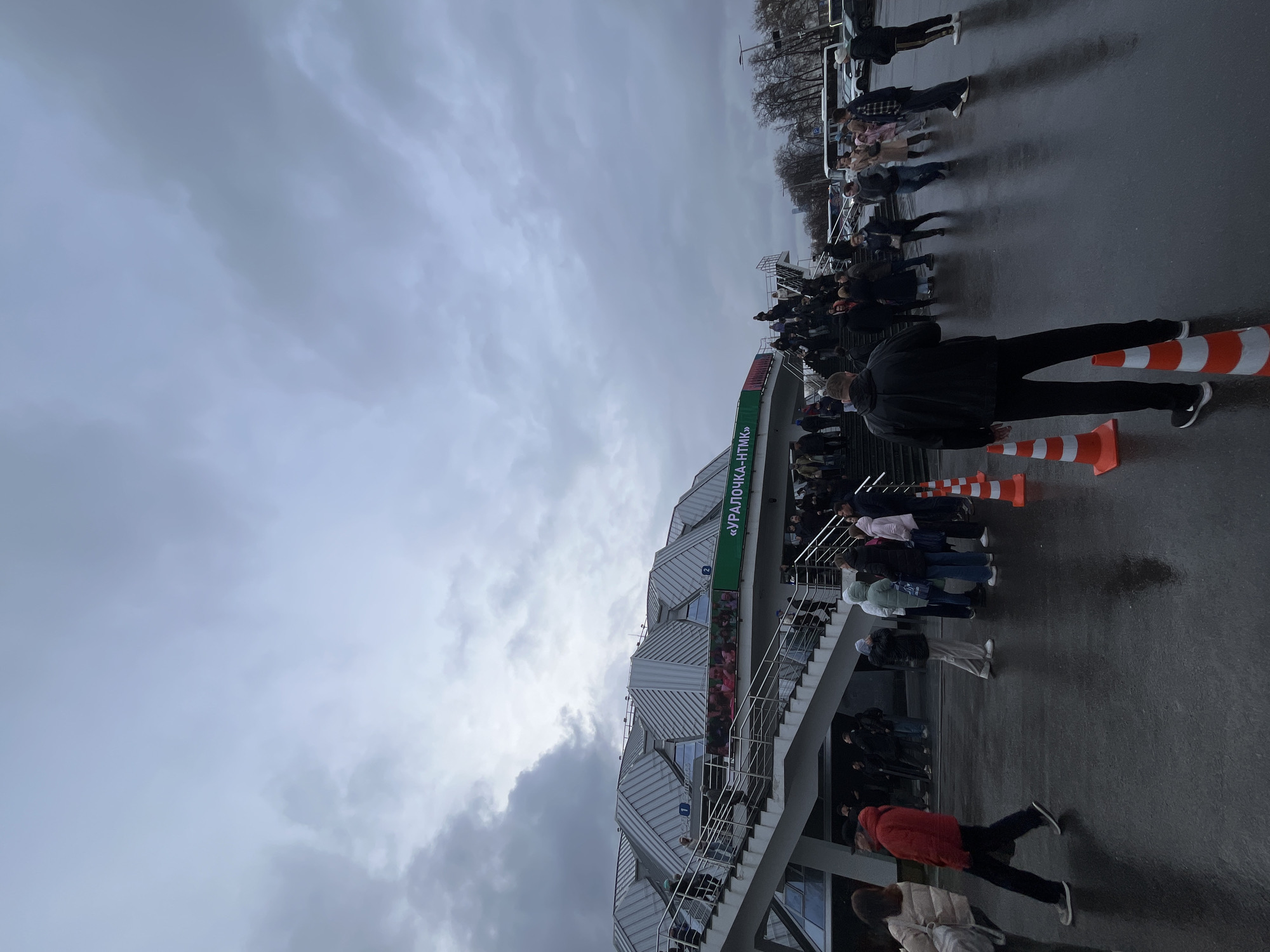 The image depicts a crowd of people queued near a stadium, with an architectural style of modern metal and glass elements. Signage on the building reads "УРАЛОЧКА-НТМК". Orange and white cones are visible, possibly used for crowd control. The ground appears wet, reflecting the grey overcast sky. The scene seems to be an exterior location, possibly the entrance to a stadium or event venue. The image is potentially taken in Russia, given the Cyrillic text on the stadium. The stadium's architecture and signage match the "Uralochka Stadium" in Moscow, Russia.