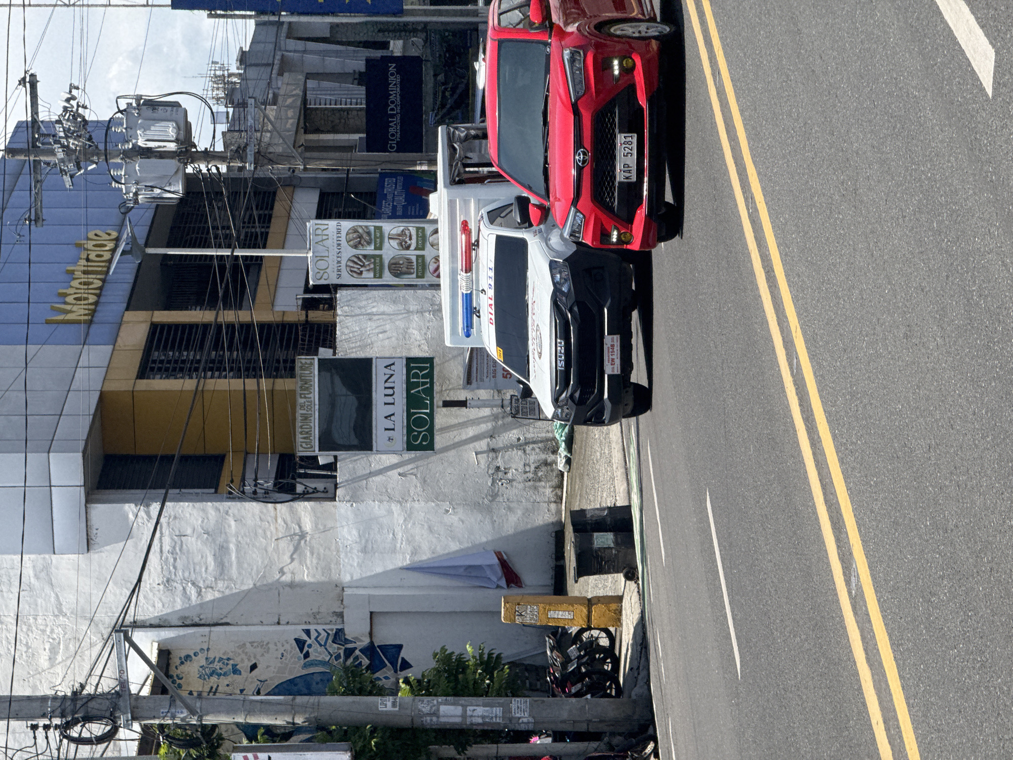 An urban street scene in Davao City, Philippines, is captured during the daytime under clear, sunny weather. Two vehicles are prominently featured: a red SUV/crossover, possibly a Toyota Raize, with the license plate KAP 5281, and a white pickup truck, an Isuzu D-Max, equipped with a red and blue emergency light bar and displaying "DIAL 911" and license plate EW 1948. Both vehicles are stopped or parked near the right side of the road, which features double yellow solid lines and a white dashed line.
On the left side of the street, multiple buildings line the sidewalk. A large blue building prominently features a "Motortrade" sign in gold lettering. Other businesses visible through various signs include "GLOBAL DOMINION FINANCING INCORPORATED" and "GIARDINI DELLE FURNITURE." Several "SOLARI" signs advertise "SERVICES OFFERED" such as "MANICURE," "NAIL ART," "PEDICURE," and "FOOT SPA," with one also mentioning "LA LUNA." Utility poles with complex, exposed wiring crisscross the frame, carrying transformers and cables. Below one building, a white wall shows signs of wear and features a mosaic-like mural with blue and white patterns. Motorbikes are parked on the sidewalk behind yellow concrete barriers. The bright sunlight creates sharp shadows, indicating midday conditions.
