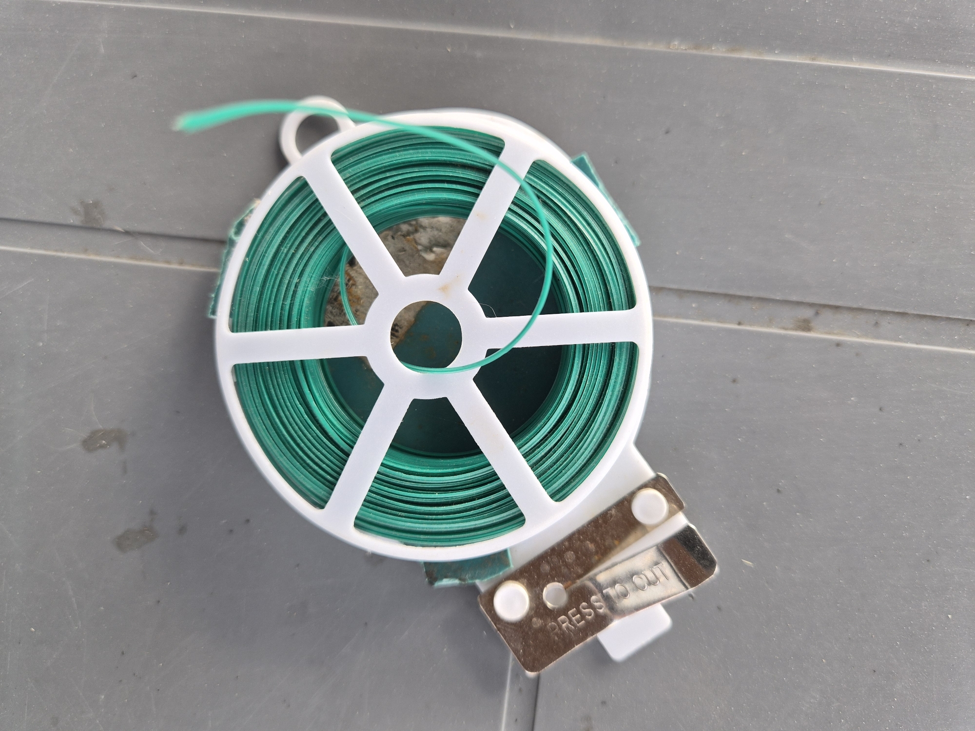 The image shows a close-up, top-down view of a roll of green garden tie wire on a gray, textured surface. The wire is wound tightly around a white plastic spool with a white plastic frame. A small metal lever with the embossed text "PRESS TO CUT" is attached to the bottom of the spool, indicating it's a cutting mechanism for the wire. A single strand of the green wire is visible emerging from the roll and looping towards the top left of the frame. The surface it rests on appears to be outdoors, possibly a deck or patio, given its ribbed texture and a few smudges that could be dirt or water stains. There are no people visible in the image. The lighting suggests it could be daytime, but no specific time or weather conditions can be determined. The context of Lenina, Russia is provided, but there are no visual cues within the image to confirm this specific location. The overall scene is utilitarian, focused on the object and its function.