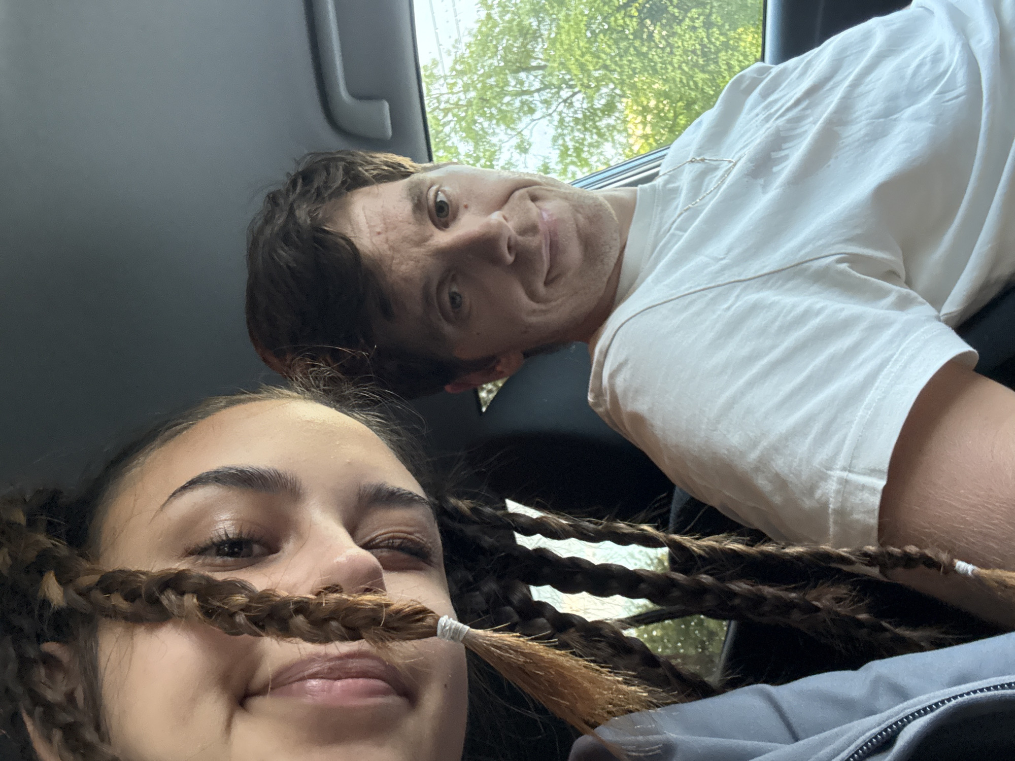 A selfie from inside a car shows two people. A young woman in the foreground, smiling slightly, has her braid across her face like a mustache. A man is in the background with a playful expression. Outside the car, green trees and foliage are visible through the car window. The location cannot be determined as Sofia, Bulgaria from the visual information in the image.