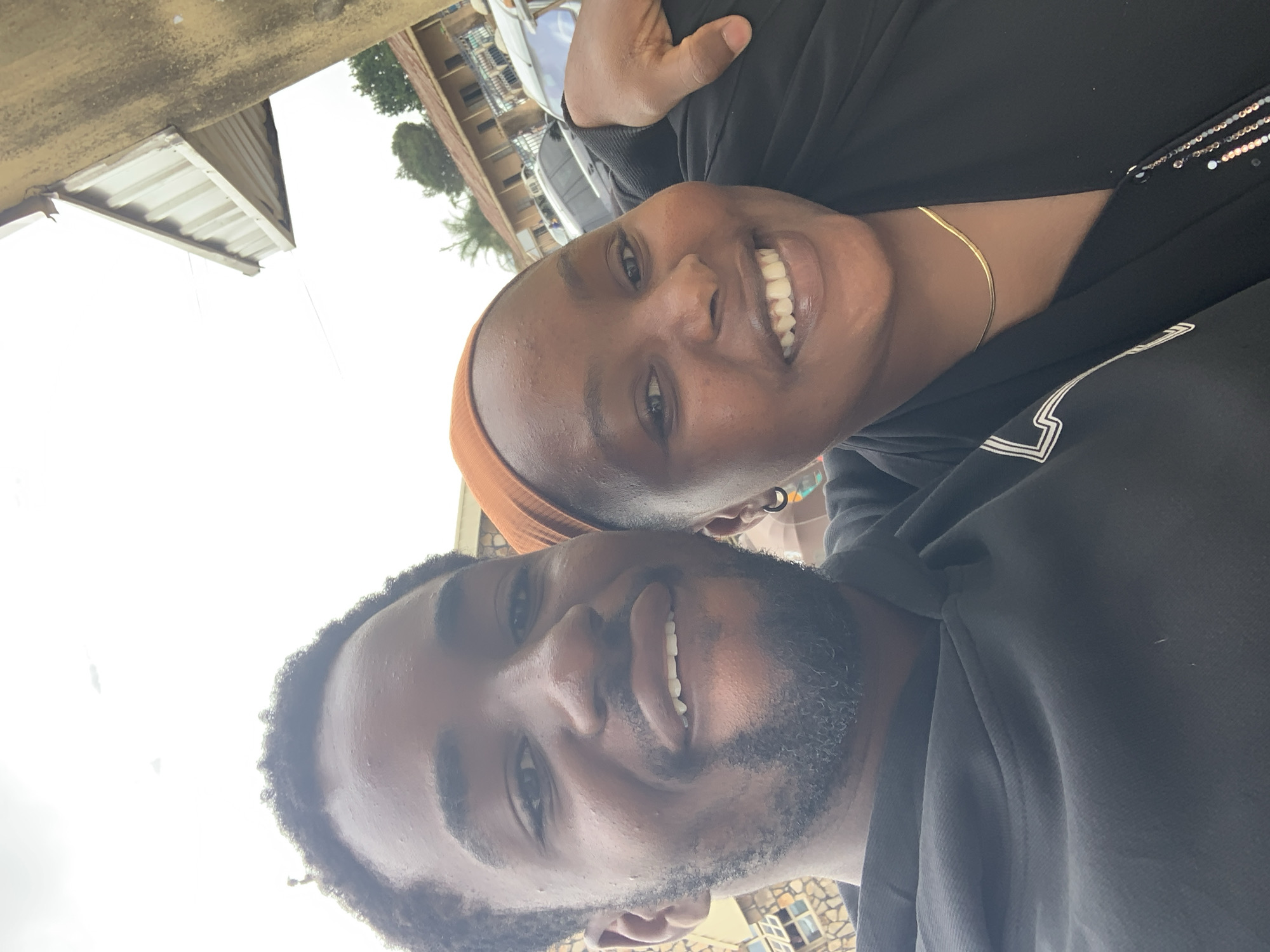 A candid outdoor portrait captures two individuals, a male and a female, in the city of Jos, Nigeria. The male, positioned on the left, has dark skin, short dark curly hair, and a full beard, wearing a dark hooded sweatshirt. He is smiling, revealing his upper and lower teeth. The female, on the right, also has dark skin, an orange headwrap, and a dark top adorned with a gold serpentine necklace and a black hoop earring in her visible left ear. She is smiling broadly, revealing her teeth, with her left arm resting over the male's right shoulder. The background features an overcast bright sky above various urban structures, including a light-brown building with a corrugated metal roof, another building with patterned stone or tile work, an overhead corrugated metal awning, and several partially visible parked vehicles. Green foliage and trees are visible between the structures, and a thin overhead wire is present in the upper left of the frame.