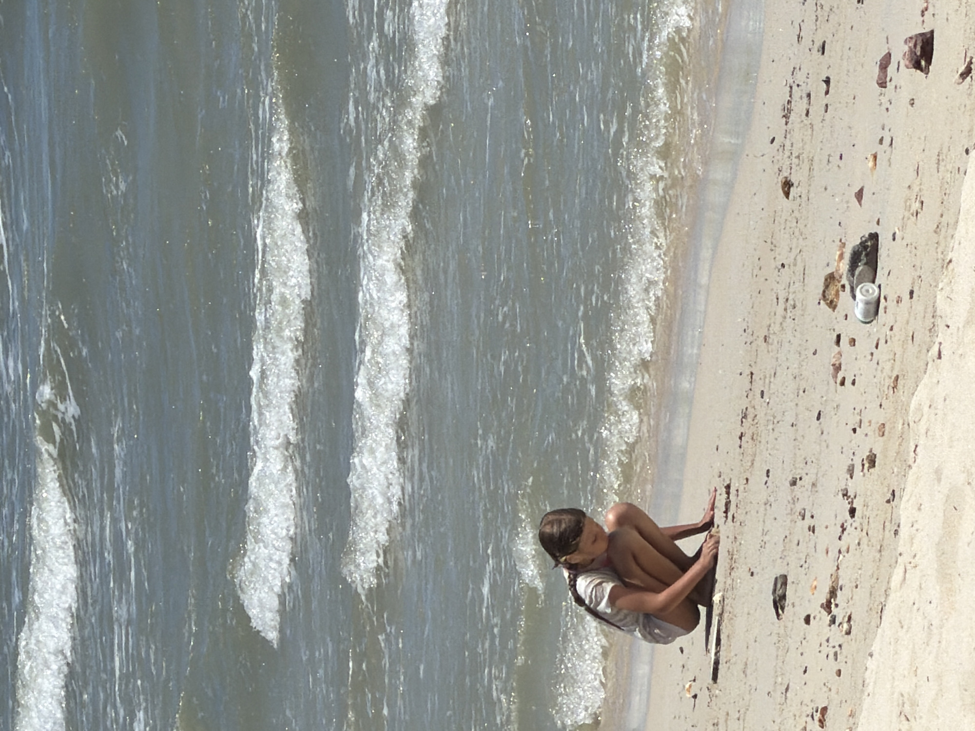 The image depicts a young girl sitting on a sandy beach next to the ocean. She is wearing a white t-shirt and shorts. Her legs are tucked under her, and her arms are resting on her knees. The girl has her hair in a braid that hangs down her back. She is looking down at her hands, which are resting on the sand.
The beach is sandy with scattered rocks and pebbles. The ocean has gentle waves rolling in, with white foam at the crests. The water is a muted blue-green color.
In the foreground, near the girl, there is a small white cylindrical object, possibly a bottle or container. There are also a few darker rocks and debris on the sand.
The scene appears to be outdoors during the daytime, with sunlight illuminating the sand and water. The weather seems to be pleasant, as suggested by the calm sea and the girl's attire. The overall mood is peaceful and contemplative.
The location is identified as Baan Tai, Thailand, which is a beach area. This context suggests a tropical or subtropical environment.
No visible text is present in the image.