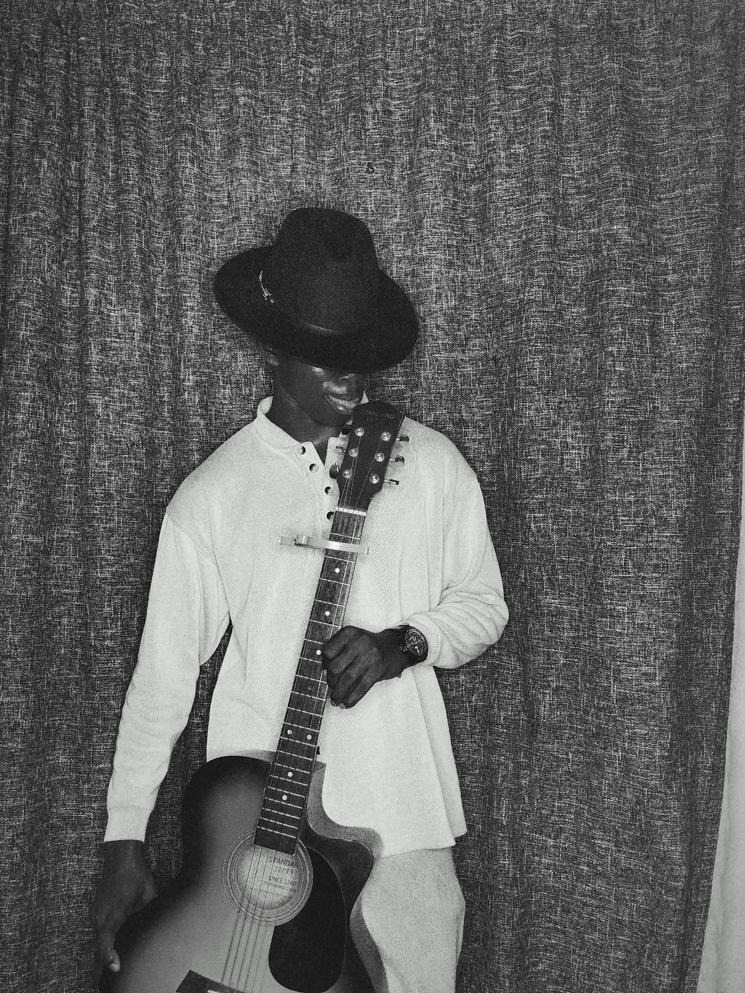 A man in a hat stands holding a guitar in front of a patterned curtain. He wears a long-sleeved shirt and a wristwatch. The guitar has a capo on the neck. The city and location are Unknown, Unknown.