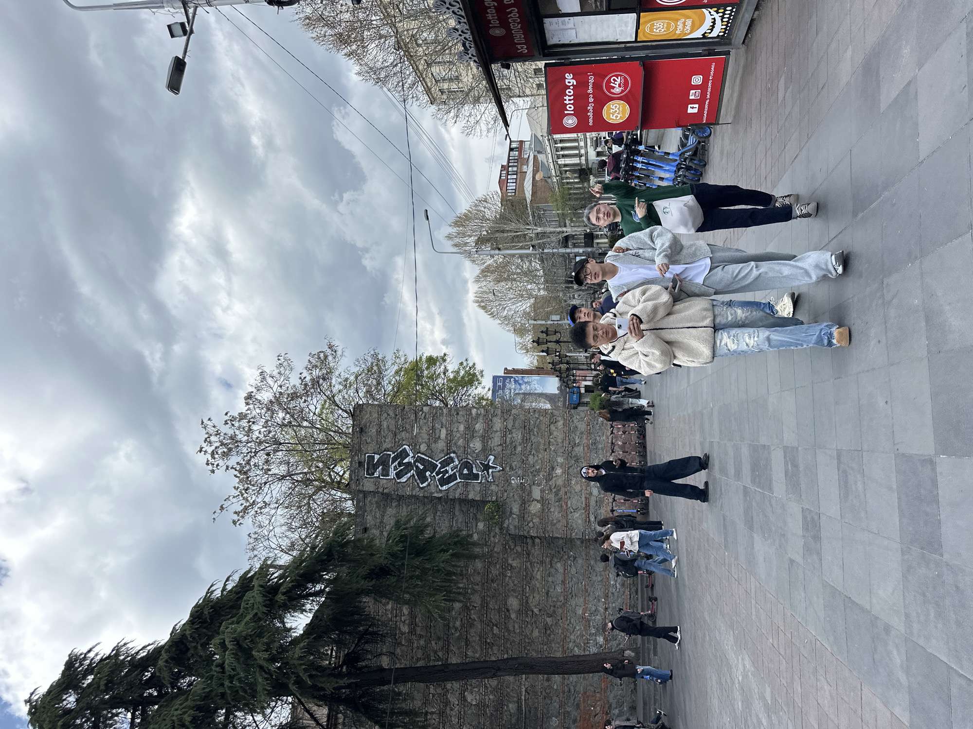 The image captures a street scene in Tbilisi, Georgia. The sky is overcast with grey clouds. Three young Asian people stand close together posing for a photo in the foreground. Behind them is a stone wall bearing graffiti, and a tree partially obscures the wall. People are walking casually on the sidewalk behind the wall. To the right of the posed group, several people are also walking. In the upper right is a red Lotto.ge sign. Blue scooters are parked nearby.