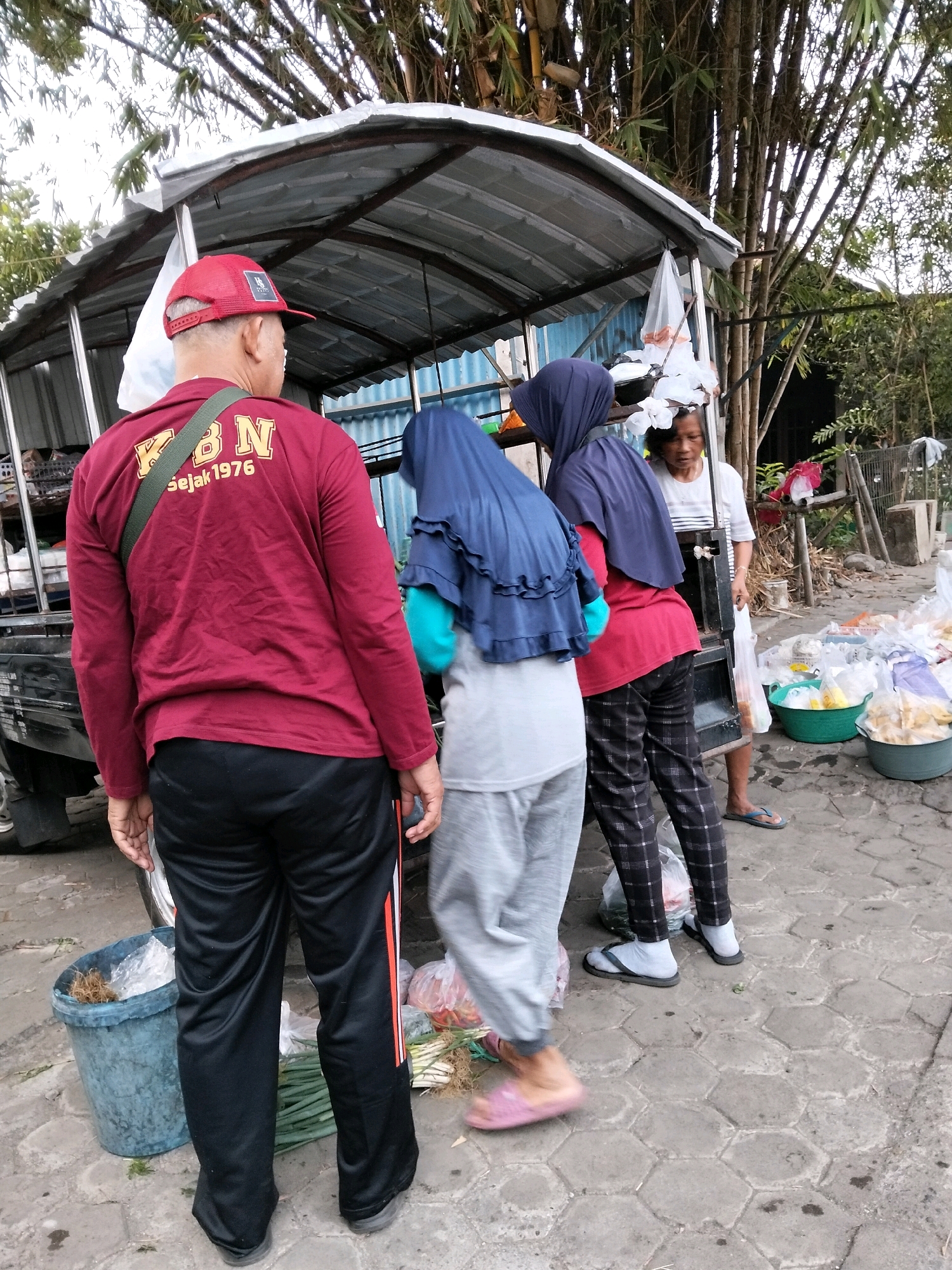 A group of people stand around a mobile market stall under a canopy, possibly in Pangenjurutengah, Indonesia. A man in a red cap and a maroon long-sleeved shirt with yellow text "KBN Sejak 1976" is in the foreground, facing away from the camera. He wears black track pants with red and white stripes. Beside him, two women wearing hijabs are interacting with the stall. One woman in a dark blue hijab wears a light blue shirt and grey pants, and a second woman in a navy hijab wears a red shirt and patterned pants. A woman in a white shirt stands further back near the stall. The stall itself is a simple structure with a corrugated metal roof. In the background, a truck is partially visible. The ground is paved with hexagonal stones. Various goods are displayed in plastic bags and buckets around the stall, including what appear to be produce like green onions and possibly fruits or other food items in the green buckets. Bamboo trees are visible in the background, suggesting a natural or semi-rural setting.
