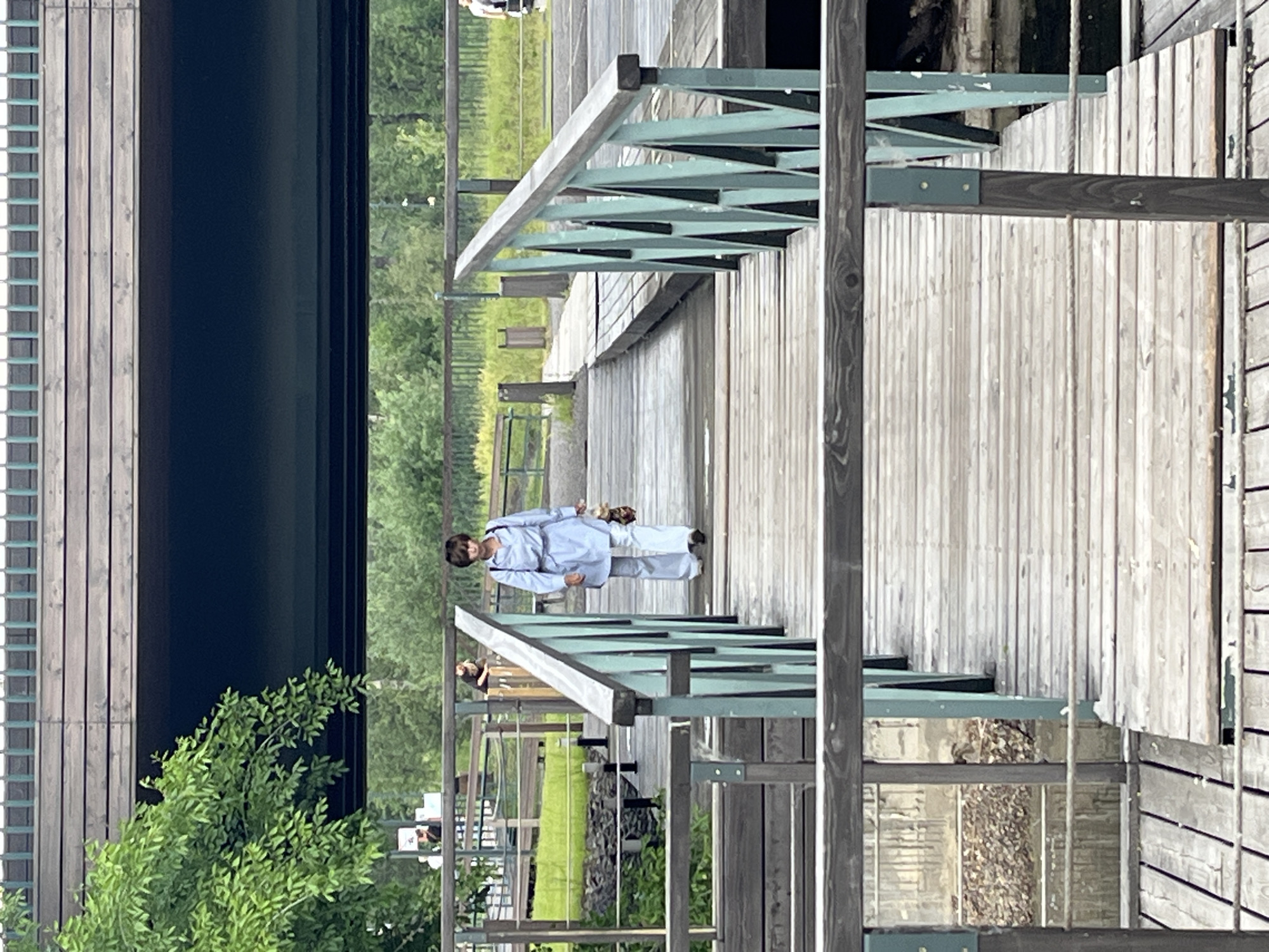 A woman walks on a wooden boardwalk under a large elevated structure in Krasnoyarsk, Russia. The woman is wearing a light blue collared shirt and matching wide-leg pants, carrying a small, patterned bag. The boardwalk is made of weathered wooden planks and has green metal railings. In the background, trees and other vegetation are visible. The elevated structure appears to be a bridge or overpass, with a dark underside and wooden planks on top.