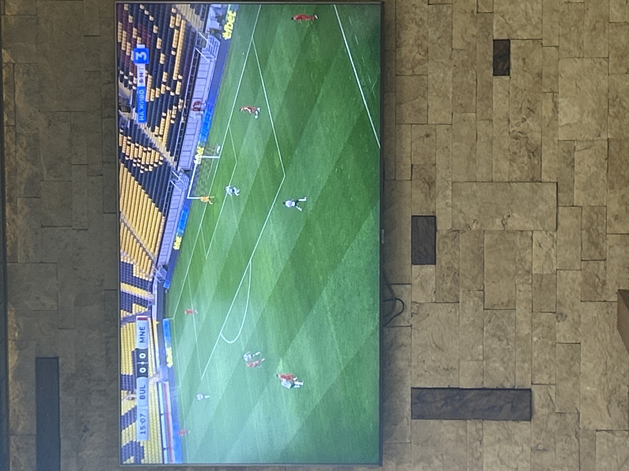 The media file displays a television screen showing a live football (soccer) match, mounted on a textured wall composed of irregular stone or tile blocks in shades of light brown and grey. Darker, rectangular blocks are integrated into the wall's design. A black cable is visible on the right side, behind the TV.
The television screen features a green football pitch with several players in various colored kits, including white, red, and dark blue/black. A player in a yellow/orange kit, likely a goalkeeper, is visible near one of the goalposts. Portions of the stadium stands with yellow and dark-colored seating are visible in the background.
Visible text on the screen includes a scoreboard in the lower left corner showing "BUL 0 0 MNE," indicating a match between Bulgaria and Montenegro with a current score of 0-0. Below the score, "15:07" is displayed, signifying 15 minutes and 7 seconds into the game. In the upper left corner, a blue banner reads "НА ЖИВО" (meaning "LIVE" in Bulgarian) and "БНТ 3," identifying the broadcast channel as BNT 3. Advertising hoardings around the pitch show the "efbet" logo. The match appears to be played during the daytime or under bright stadium lights. The overall setting is consistent with a sporting event being broadcast in Bulgaria, fitting the provided location context of Kazanlak, Bulgaria.