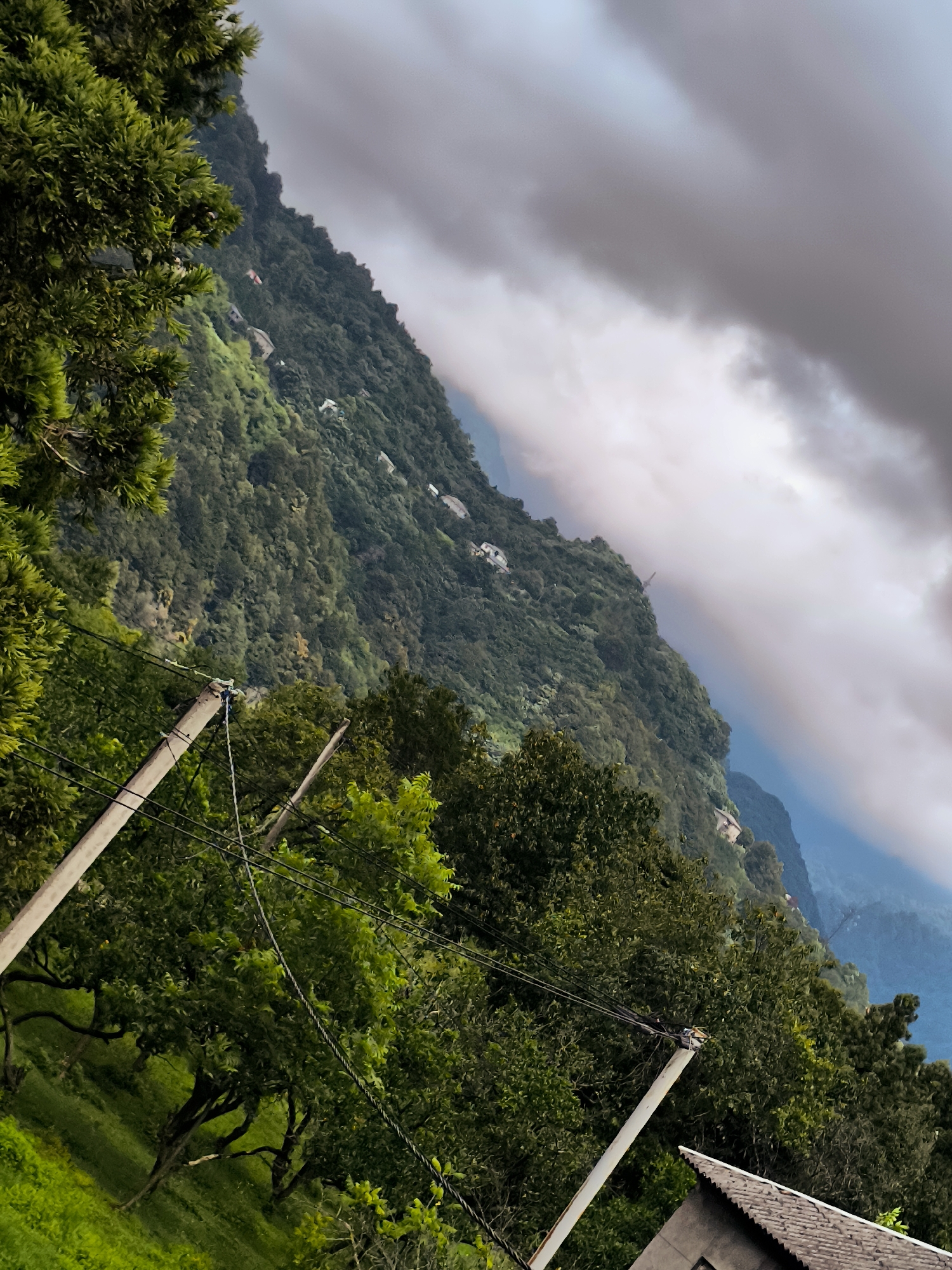 The image depicts a mountain slope covered in lush green trees, with scattered houses visible on the mountainside. A cloudy sky looms overhead, with patches of light breaking through. In the foreground, power lines are strung between two utility poles, with a portion of a tiled roof visible in the lower right corner. The scene is captured at a diagonal angle, giving a sense of depth and perspective. The specific location in Batumi, Georgia, is not identifiable from the image itself.