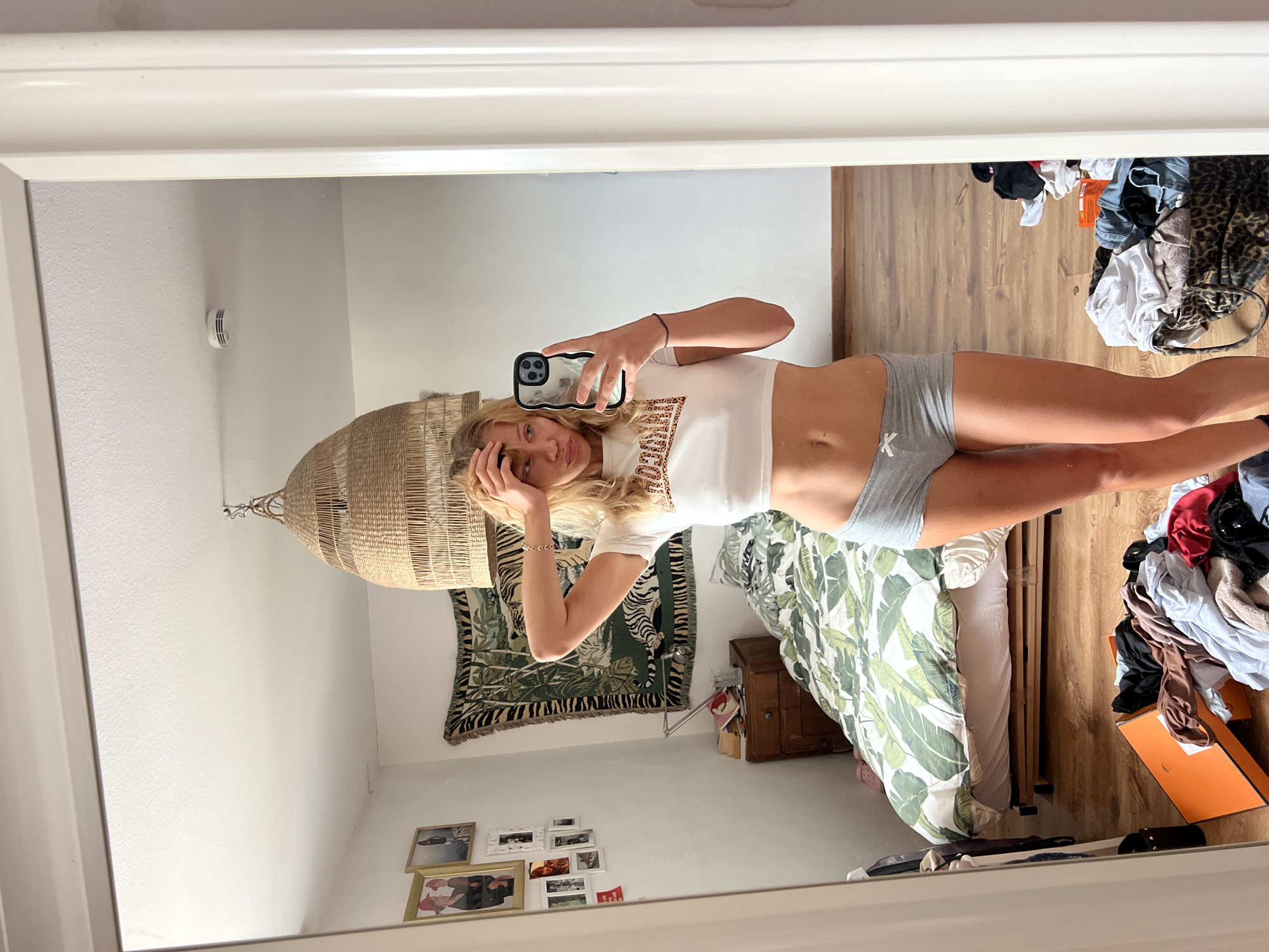 A young woman with blonde hair is taking a mirror selfie in a bedroom. She is wearing a white crop top with a leopard print design and grey athletic shorts. Her right hand is touching her head, and her left hand is holding her phone. The room has a wooden floor and white walls. A large woven pendant light hangs from the ceiling. There is a bed with a duvet featuring a tropical leaf pattern. A wooden dresser is beside the bed, and a pile of clothes is on the floor near the dresser. A tapestry with a zebra pattern hangs on the wall. Several framed pictures are on the wall. The scene appears to be indoors, possibly in a residential setting. The lighting suggests it is daytime. There is no visible text in the image.