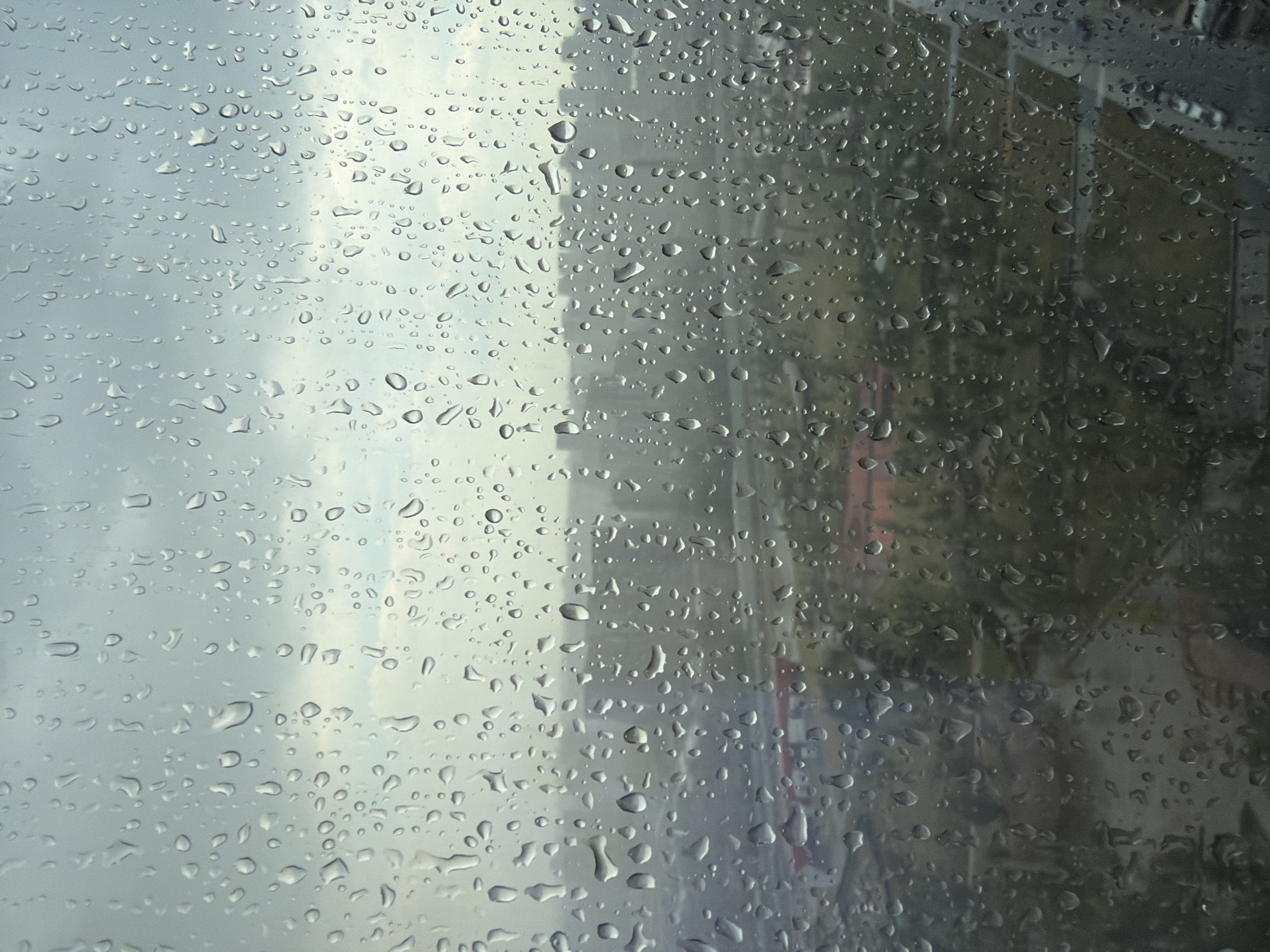 The image shows a windowpane with numerous raindrops on it, distorting the view of a city landscape outside. The cityscape is hazy and appears to be an urban environment with buildings and some greenery. The overall mood of the image is subdued and somber due to the rain and the overcast sky, typical of a cloudy day. The specific location in Moscow, Russia, cannot be determined from the provided image.
