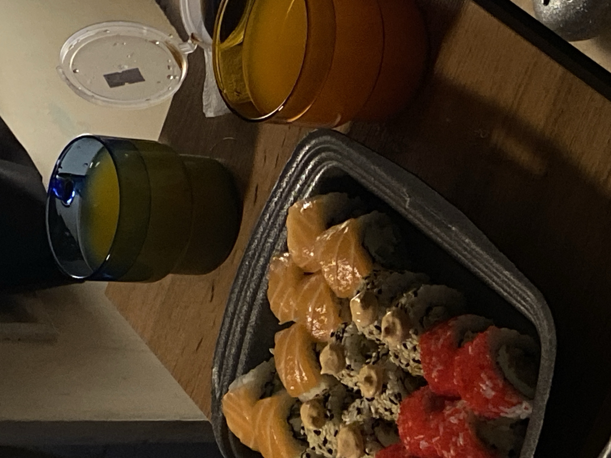 The image shows a table setting, possibly indoors. On the table, there is a black tray containing a variety of sushi rolls, including salmon nigiri and rolls with red tobiko. Beside the tray, there are two glasses of orange juice, one in a green-tinted glass and another in a brown-tinted glass, with a translucent lid nearby. The background suggests a simple interior with wood-like surfaces. There is no information related to the location being Votkinsk, Russia.