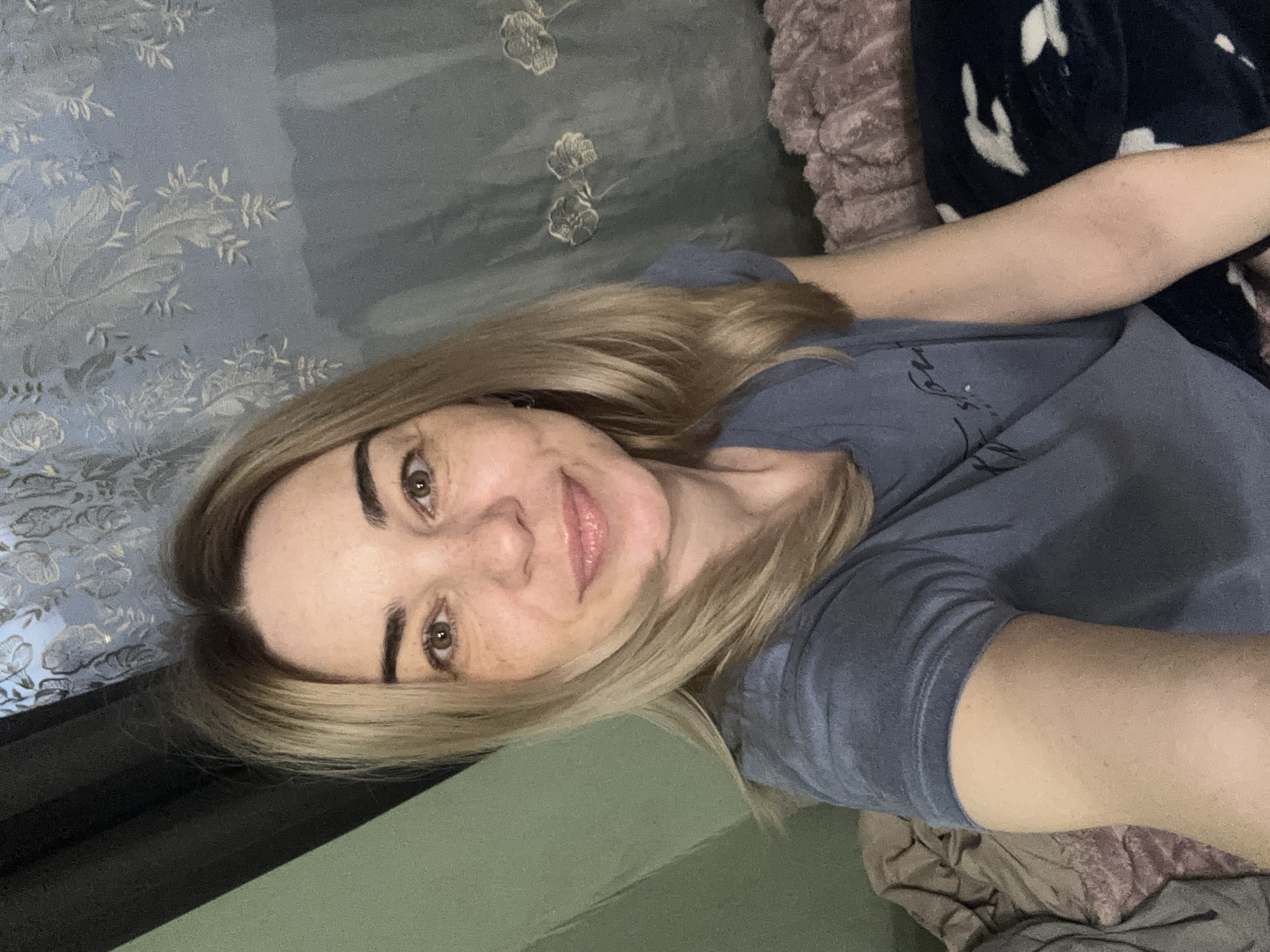 A blonde woman wearing a grey t-shirt is lying down, facing the camera. She has fair skin, light brown eyes, and is smiling slightly. The background includes a blue floral patterned curtain, a pink textured blanket, and a dark blue blanket with white spots. No information available to locate it in the city of Vsevolozhsk, Russia.