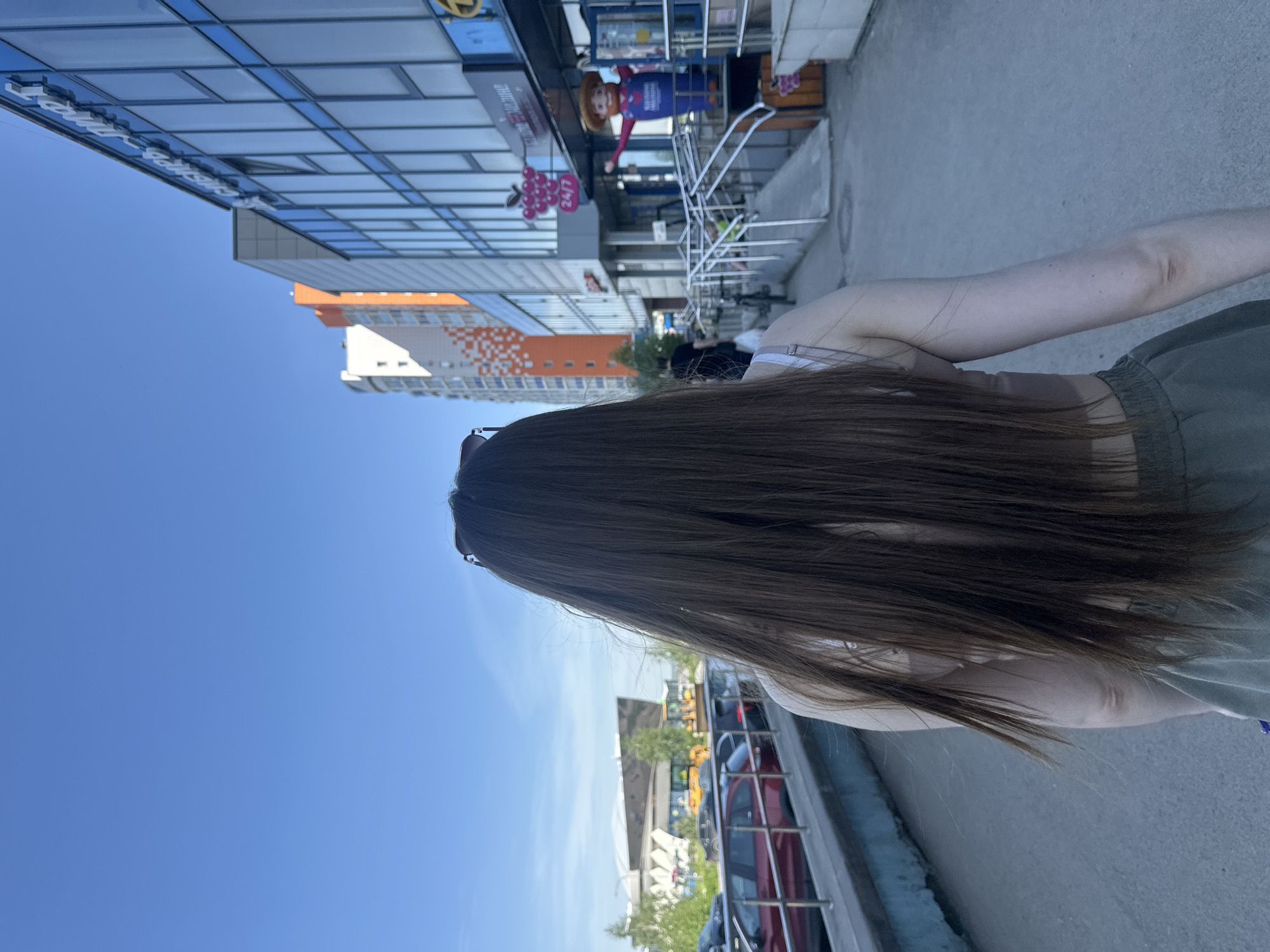 A person is depicted from behind, their long, straight dark brown hair flowing down their back. They are wearing a light-colored top and olive green cargo-style pants or shorts. Sunglasses are perched on top of their head. The setting is an urban environment on a clear, sunny day, with a bright blue sky overhead.
In the background, modern buildings line a street or plaza. To the left, a building with reflective blue glass facades prominently features white text, which appears to be "Лондон-Парк" (London Park). Further back, another multi-story building displays a pattern of orange and white sections. On the right side, near the blue glass building, a large, inflatable mascot figure, resembling a cowboy or farmer in a blue shirt and brown hat, stands next to a building entrance. A small sign shaped like purple grapes with "24/7" written on it is also visible.
The person appears to be walking or standing on a paved sidewalk or street, looking towards the buildings and distant cars. Several vehicles are parked or driving in the mid-ground, including a red car. The overall scene suggests a commercial or mixed-use area of a city during daytime.
