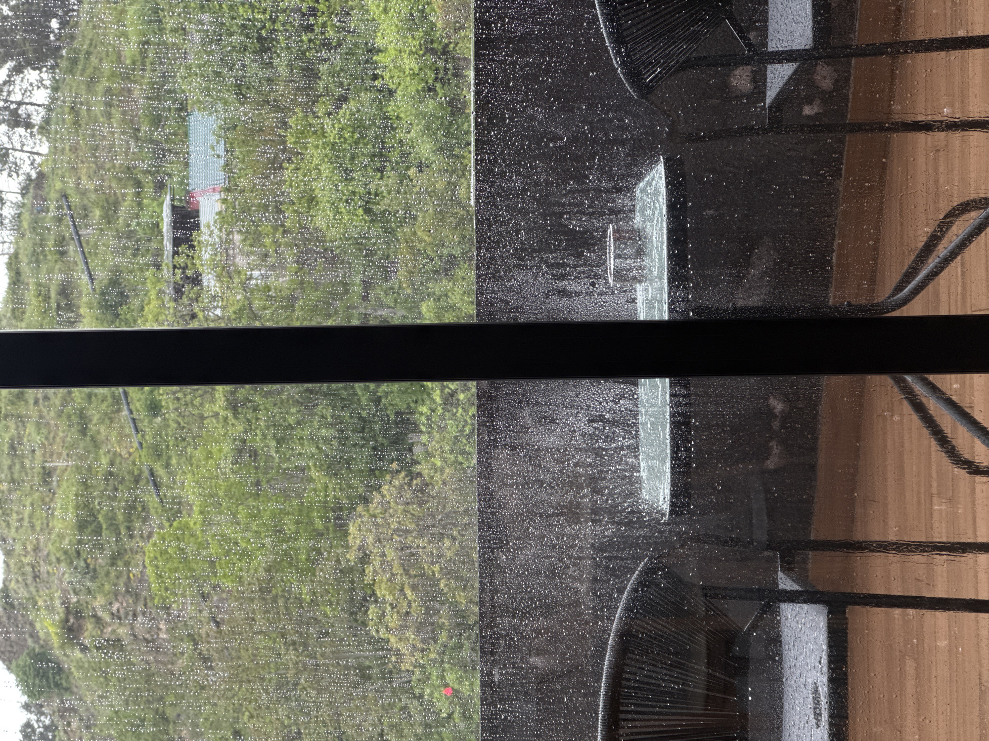 The image shows a view through a wet window, likely due to rain. Outside the window, there's a green landscape with trees and a building visible in the distance on the left side of the window. On the right side of the window, a chair is visible, with parts of the chair's frame and seat appearing out of focus. The window frame is black and runs horizontally across the center of the image. The location is described as Unknown, Unknown.