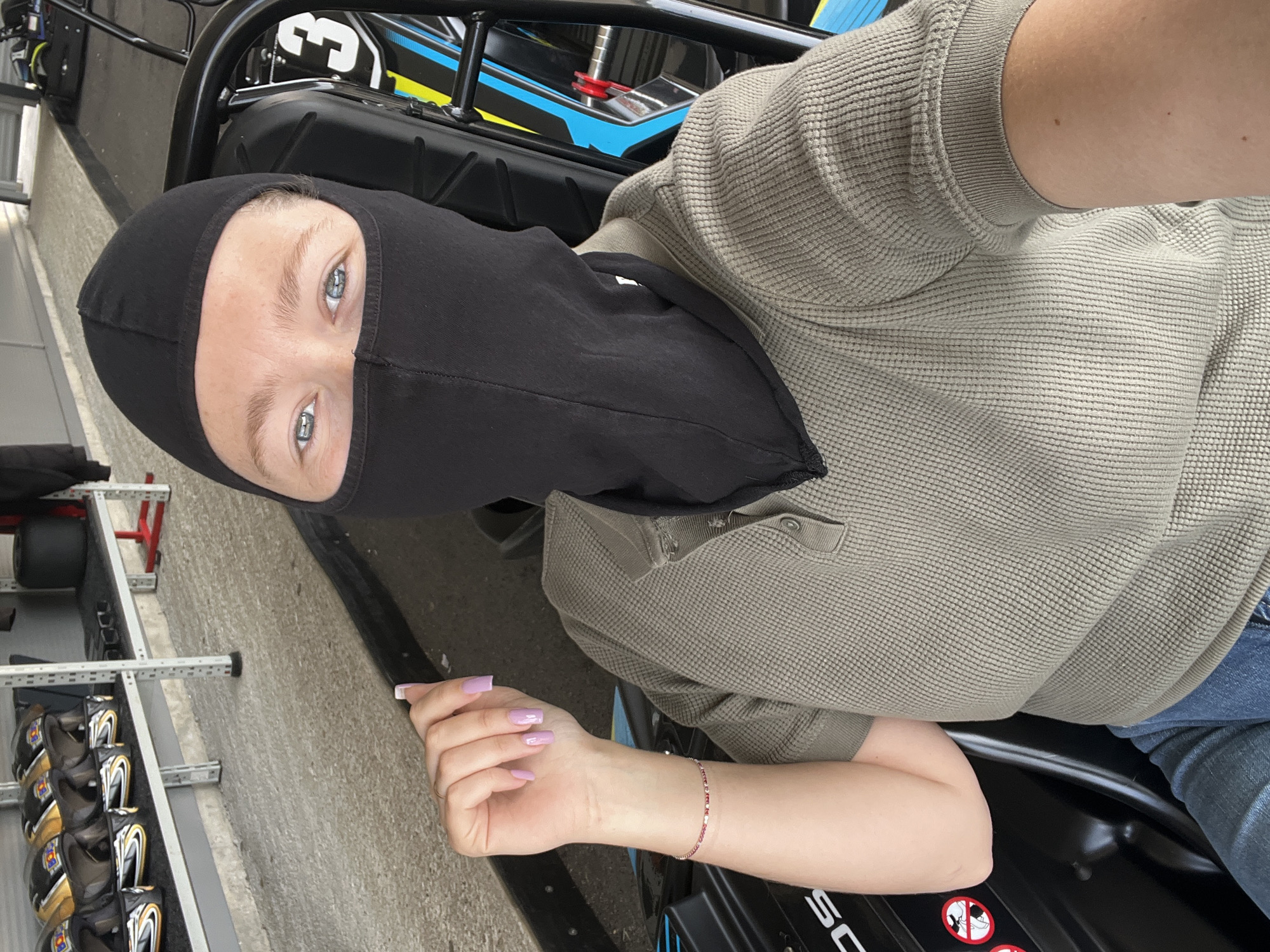 A person wearing a black balaclava and a grey waffle-knit shirt is sitting in a go-kart. The person has light blue eyes and painted fingernails. A helmet rack with helmets is visible on the left side of the image. The location is Bigor, Montenegro.