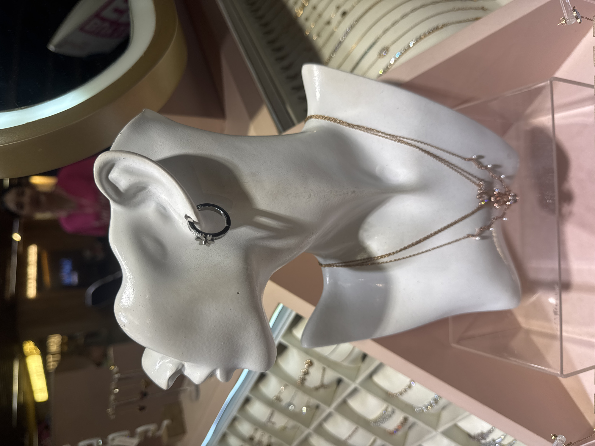The image features a white, stylized mannequin bust, resembling a head and upper torso, prominently displaying jewelry in what appears to be a retail setting. On the mannequin's left ear, a silver-toned hoop earring with a small, floral or star-shaped charm is visible. Around its neck, a delicate, multi-layered gold-toned necklace is draped, featuring small charms or embellishments on its side.
The background is softly blurred, but details suggest a jewelry store or boutique environment. To the upper left, a round mirror with a golden frame reflects blurred shapes and colors, including a faint, out-of-focus reflection of a person's face. Below and to the right of the mannequin, various trays filled with numerous pieces of jewelry, possibly earrings or smaller necklaces, are visible, further confirming the retail context. The overall lighting is artificial and warm, highlighting the jewelry and the display. There is no discernible text visible in the image. The scene is static, focused on the presentation of merchandise. The location context is Kuta Selatan, Indonesia, indicating a commercial establishment in that area.