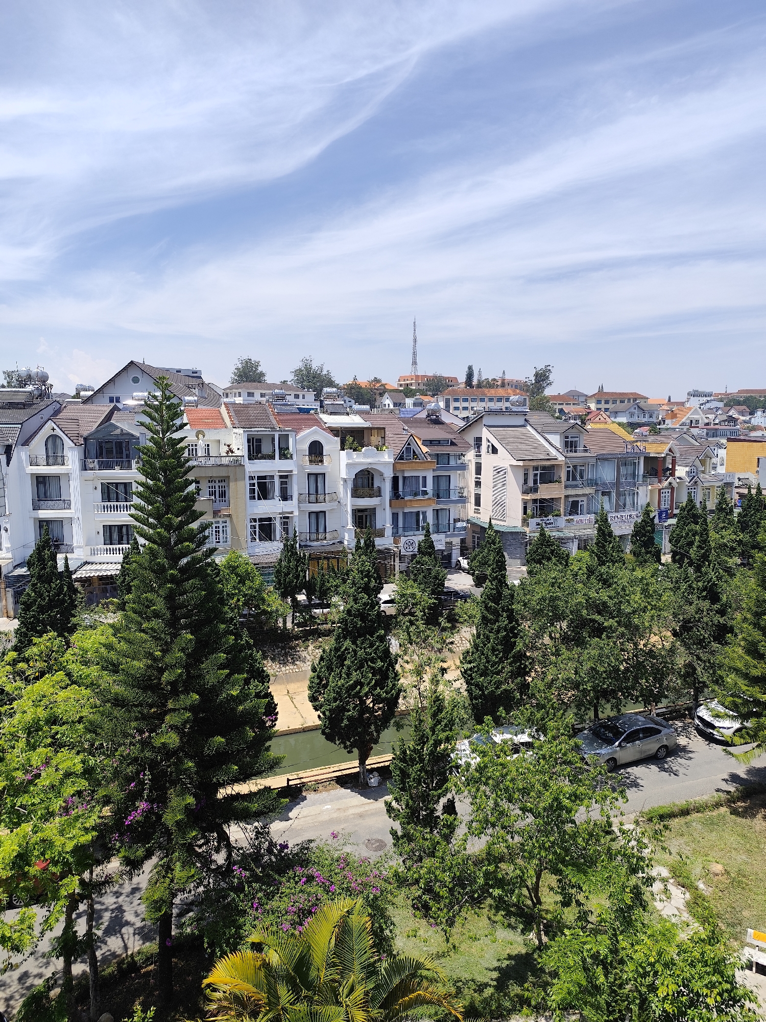 The image presents an elevated view of a section in Da Lat, Vietnam. In the foreground, verdant foliage predominates, with several mature trees. A narrow, concrete-lined canal runs horizontally across the mid-ground. A street with parked vehicles is discernible. In the background, a row of multi-story buildings lines the horizon. The architectural style suggests a European influence, with pitched roofs, balconies, and white exteriors. Distant hills and a communication tower are seen in the far background under a partly cloudy blue sky.