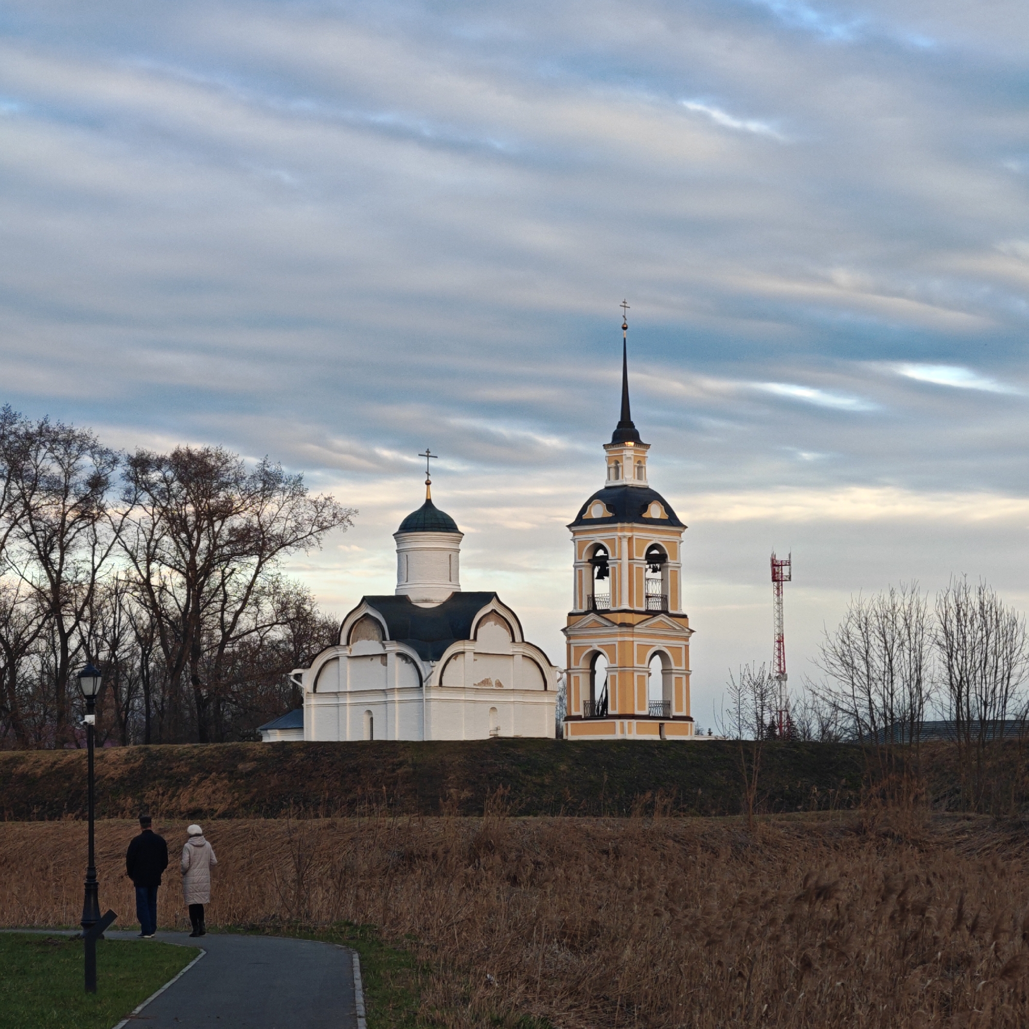 The image depicts a couple walking on a path in front of a church complex on a slightly elevated area. The church is composed of a white building with a dark roof and a separate yellow and white bell tower with a black spire and cross. Behind the church, there's a cell phone tower. The foreground is dominated by dry, brown vegetation. A black lamppost stands to the left of the couple. The sky is overcast with striated clouds. This is likely a scene within the city of Suzdal, Russia, known for its historical churches.