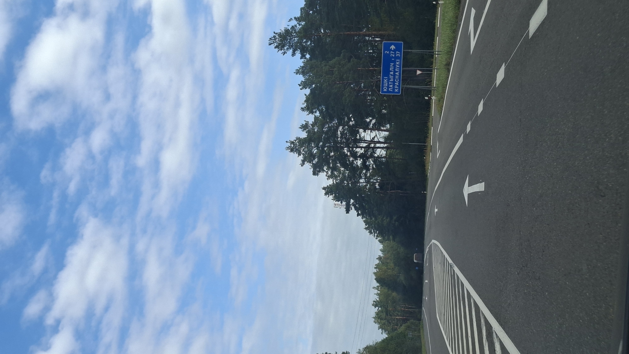 The image is a vertical shot taken from a vehicle on a paved road in what appears to be a rural area or the outskirts of a city in the Лепельскі сельскі Савет, Belarus. The road has multiple lanes with clear white lane markings and arrows indicating the direction of travel. On the right side of the road, a blue directional signpost is visible, displaying place names in Belarusian: "ЮШКІ 2", "ЛАТЫГАЛІЧ 27", and "КРАСНАЛУКІ 37". Dense green pine trees line the right side of the road, creating a natural border. The sky above is a clear blue with scattered white, fluffy clouds. In the distance, a white van or truck is seen driving in the opposite direction. The overall scene depicts a travel scenario on a well-maintained road surrounded by a natural landscape.