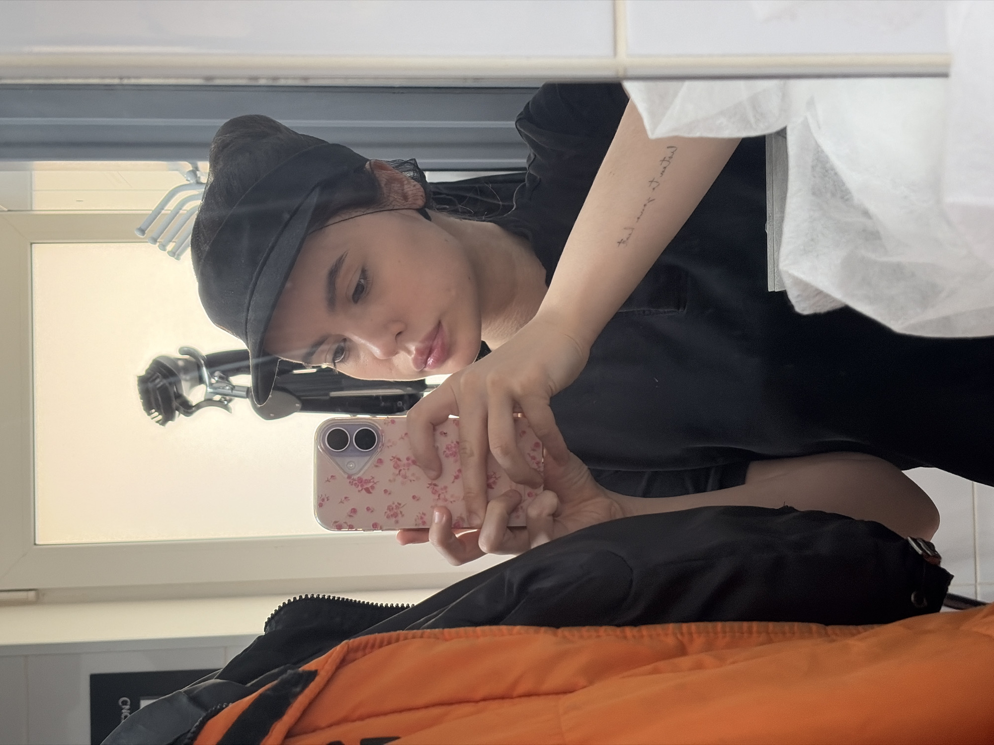 A young person with light skin, wearing a black cap and dark shirt, is taking a selfie in front of a mirror. The person is holding a pink floral patterned smartphone. Reflected in the mirror are window blinds and a white towel dispenser. The individual is sitting on an orange and black jacket. The location is Chaikovsky, Russia.