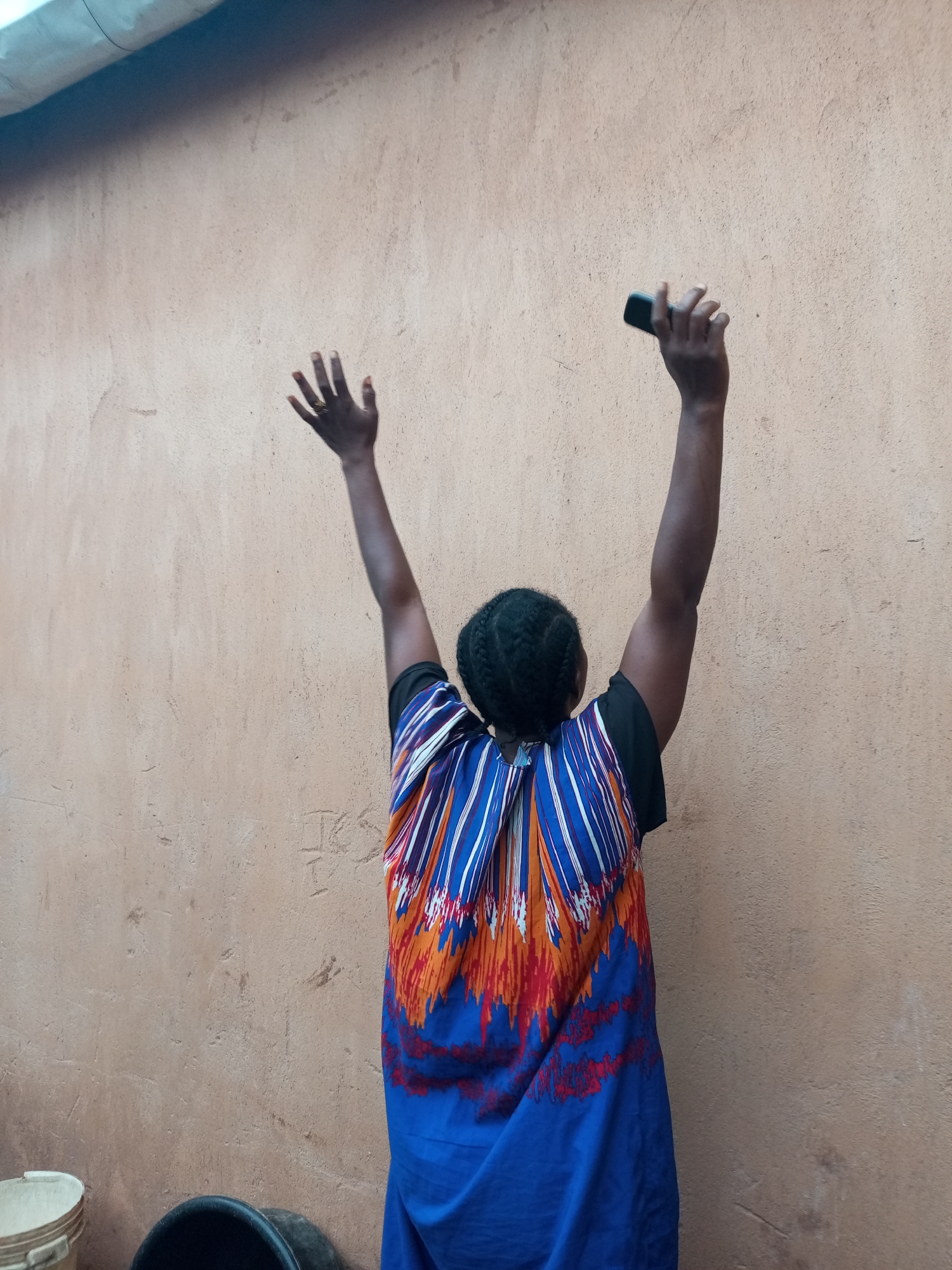 A person stands with their back to the camera, facing a light brown wall. Their arms are raised above their head, and they are holding a dark-colored object, possibly a mobile phone, in their right hand. The person is wearing a vibrant, patterned dress with blue, orange, and white vertical stripes. The dress has a black neckline. The wall appears to be made of stucco or concrete and shows signs of wear and tear, with some faint markings. In the lower left corner of the image, a white bucket and a black bucket are visible, suggesting an outdoor or utility area. The scene is set in an urban environment, likely Tempure, Nigeria, given the context. The lighting suggests it is daytime.