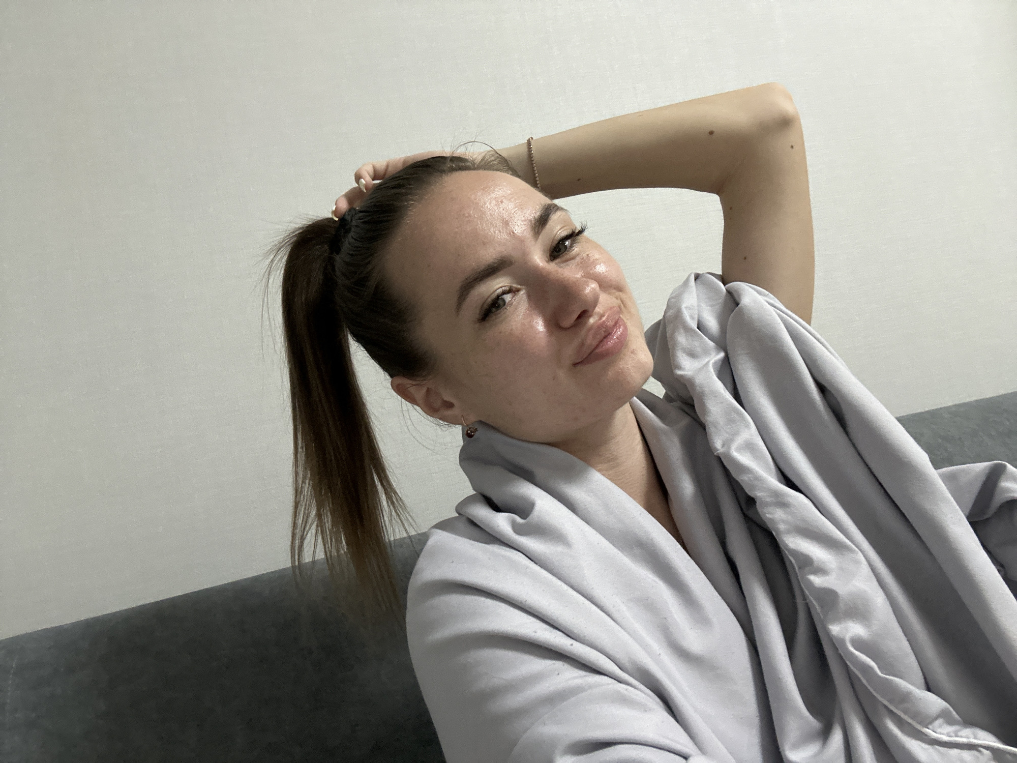 A young woman with brown hair in a ponytail poses for a selfie. She is wearing a light gray robe and has a delicate bracelet on her right arm. Her left hand is in her hair, and she is looking at the camera with a playful expression, her lips slightly puckered. She is seated on a gray couch in what appears to be a domestic setting, with a plain white wall in the background. The lighting is soft, suggesting an indoor environment.
