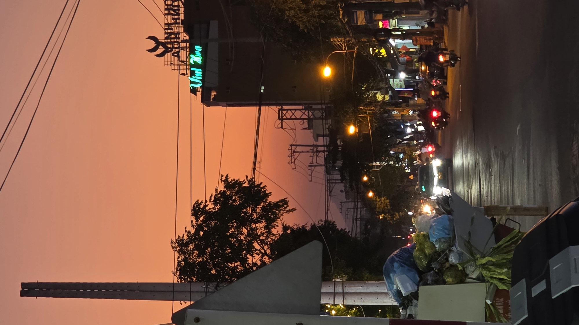 The image shows a street scene at dusk in Bắc Nha Trang, Vietnam. The sky is a warm orange color. On the left, electrical lines and a building with a green neon sign that reads "Vinh Hoa" are visible. A tree partially obscures the view. In the center, a power grid is visible.
On the right, a street with parked motorbikes and streetlights illuminates the scene. Several motorbikes are in motion on the road, identifiable by their red tail lights. In the foreground, a pile of garbage bags and debris is visible, partially blocking the view. A small portion of a vehicle is also visible in the foreground.
