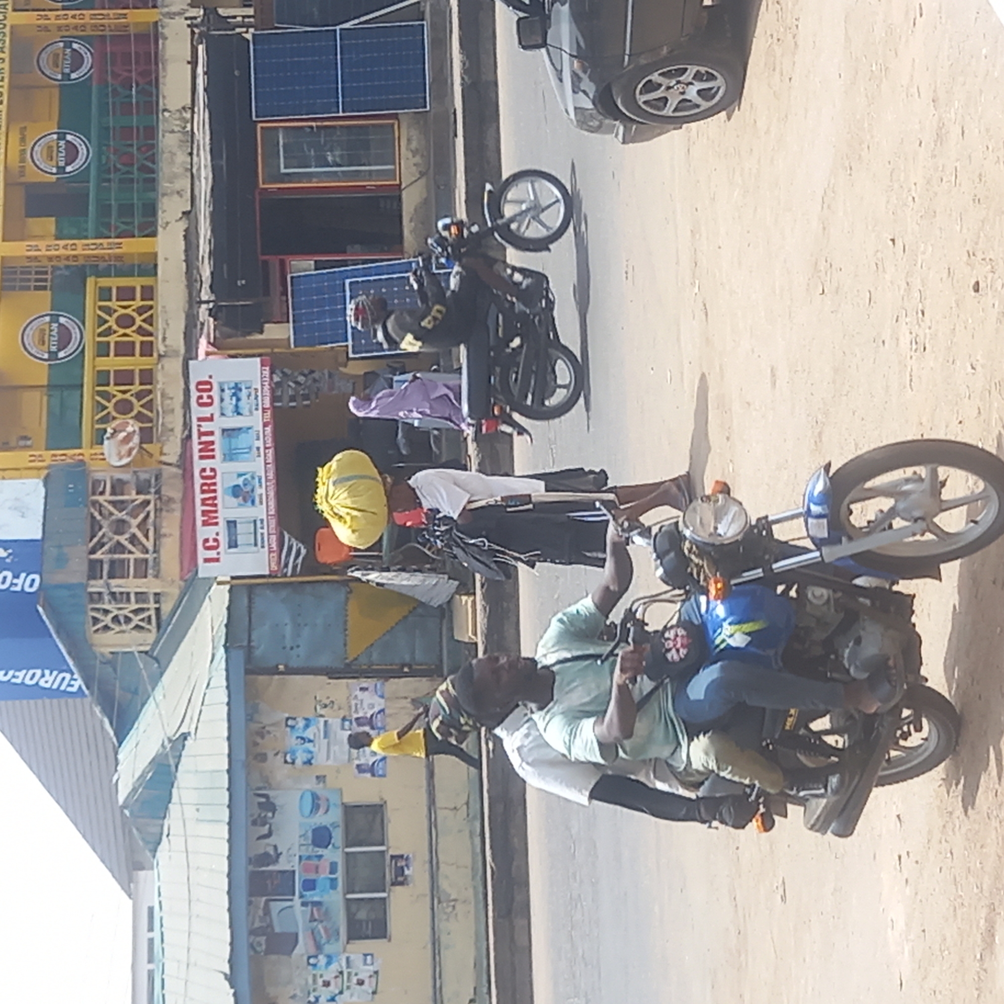 This image depicts a bustling street scene in what appears to be an urban or semi-urban area, consistent with Dadi Riba, Nigeria. The bright sunlight and strong shadows suggest it is midday.
Several people are present, primarily men. In the immediate foreground, two men are on a blue and black motorcycle, one driving and the other riding pillion. Further back, a man stands beside another dark-colored motorcycle, balancing a large yellow sack or bag on his head. Another individual is visible on a different motorcycle in the mid-ground. A black car is partially seen parked on the right side of the unpaved or dusty road.
The setting includes commercial buildings lining the street. Notably, several blue solar panels are visible on the rooftops and walls of these structures. Various goods are displayed outside shops. Prominent text on a sign reads "I.C. MARC INT'L CO." with contact details: "OFFICE LAGOS STREET SOKOTO ROAD KADUNA. TEL: 08039643282". Other visible text includes "EUROFO", "OFO", and multiple instances of "RTEAN" on yellow and green crates, likely indicating the Road Transport Employers' Association of Nigeria. The scene conveys a sense of everyday life and commerce in a busy roadside environment.
