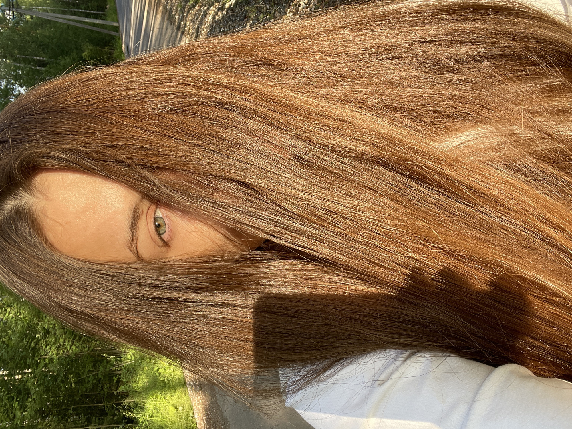 A young woman with long, auburn hair partially covers her face in this outdoor, close-up, medium-shot photograph. She is wearing a white hooded garment. Her visible eye is green, and her skin is illuminated by the warm light of the setting sun. Her hair is the dominant element, falling forward and obscuring most of her face. The background shows a gravelly road or path, lush green vegetation, and trees, suggesting a rural or semi-rural setting, possibly on the outskirts of Заречный, Russia. A shadow of a rectangular object, likely a phone, is cast on her hair.