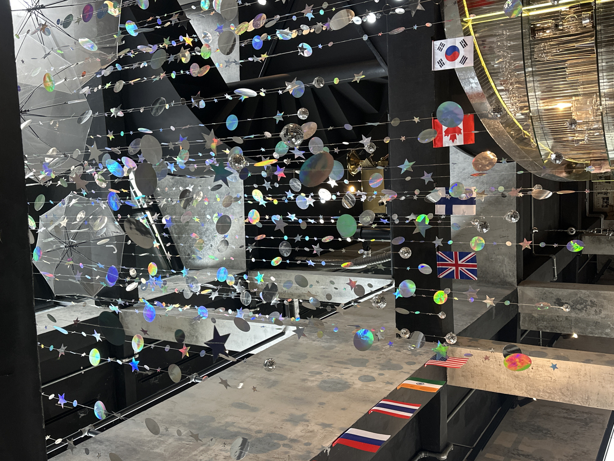 The image shows an indoor space decorated with hanging iridescent star and circle ornaments, as well as several national flags. The flags visible include Thailand, India, the United States, the United Kingdom, Finland, Canada, and South Korea. The decor hangs from the ceiling, which is dark and industrial-looking with exposed beams. In the background, a spiral staircase is visible, along with concrete walls and industrial lighting. A bar area with glassware is also present in the lower right portion of the image. The scene appears to be a restaurant or bar, likely in Đà Nẵng, Vietnam, given the international flags displayed. There are no people visible in the image.