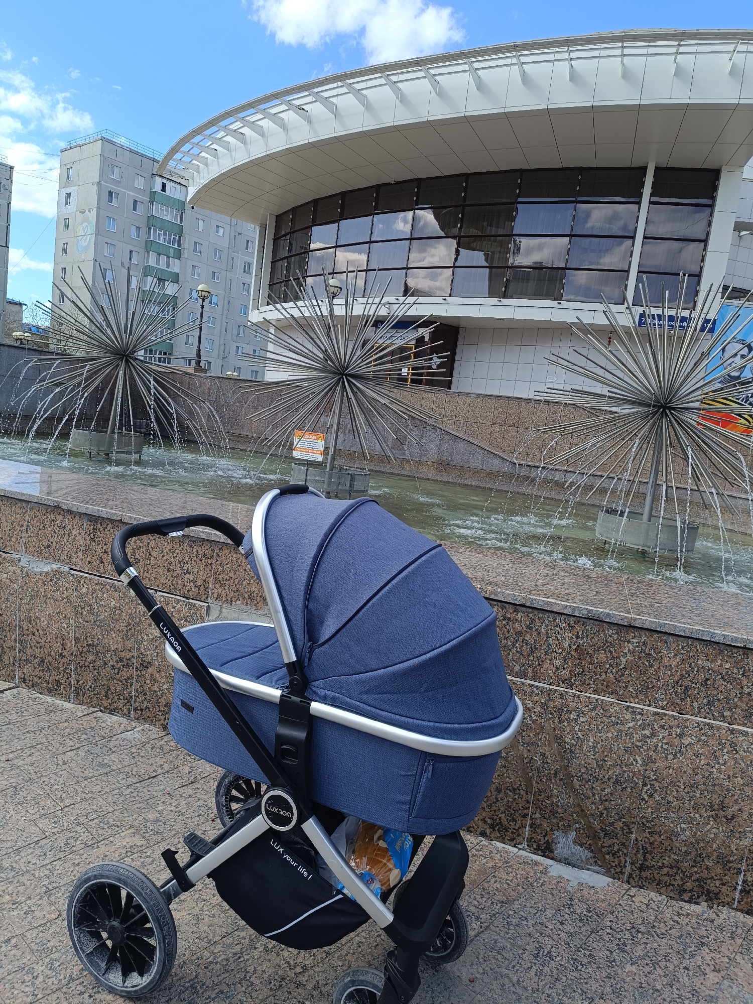 The image presents an urban outdoor scene in Tobolsk, Russia, under a bright, partially cloudy sky. In the foreground, a blue and silver baby stroller is prominently featured, parked on a paved sidewalk. The stroller's frame bears the brand name "LUXMOM," and its storage basket displays the slogan "LUX your life!"; some packaged goods are visible inside the basket.
Behind the stroller, a decorative fountain is active, with water cascading from two large, spherical metal sculptures composed of numerous spikes. The fountain pool contains greenish water and is bordered by a wall with a speckled, granite-like texture.
In the midground and background, various buildings define the setting. A modern structure with a distinctive curved, white roof and large, reflective glass windows dominates the center. These windows mirror the blue sky and white clouds. To the left and behind this modern building, older, multi-story residential apartment blocks with numerous windows are visible. One of these buildings features a light green section and a faded emblem or artwork on its side.
The sky is bright blue with scattered white clouds, indicating daytime. Visible text includes "LUXMOM" and "LUX your life!" on the stroller. Near the fountain, a sign displays Russian text, including "ВНИМАНИЕ" (Attention/Warning) and "7 м микрорайон" (7th microdistrict), providing a specific location reference within Tobolsk. The scene depicts a public area combining modern and older architectural styles.